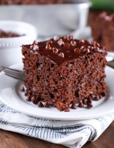 Slice of easy chocolate zucchini cake on a white plate with a pan of cake in the background. Recipe from A Kitchen Addiction