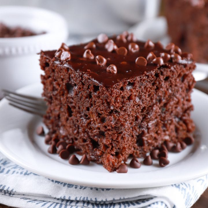 Slice of easy chocolate zucchini cake on a white plate with a pan of cake in the background. Recipe from A Kitchen Addiction