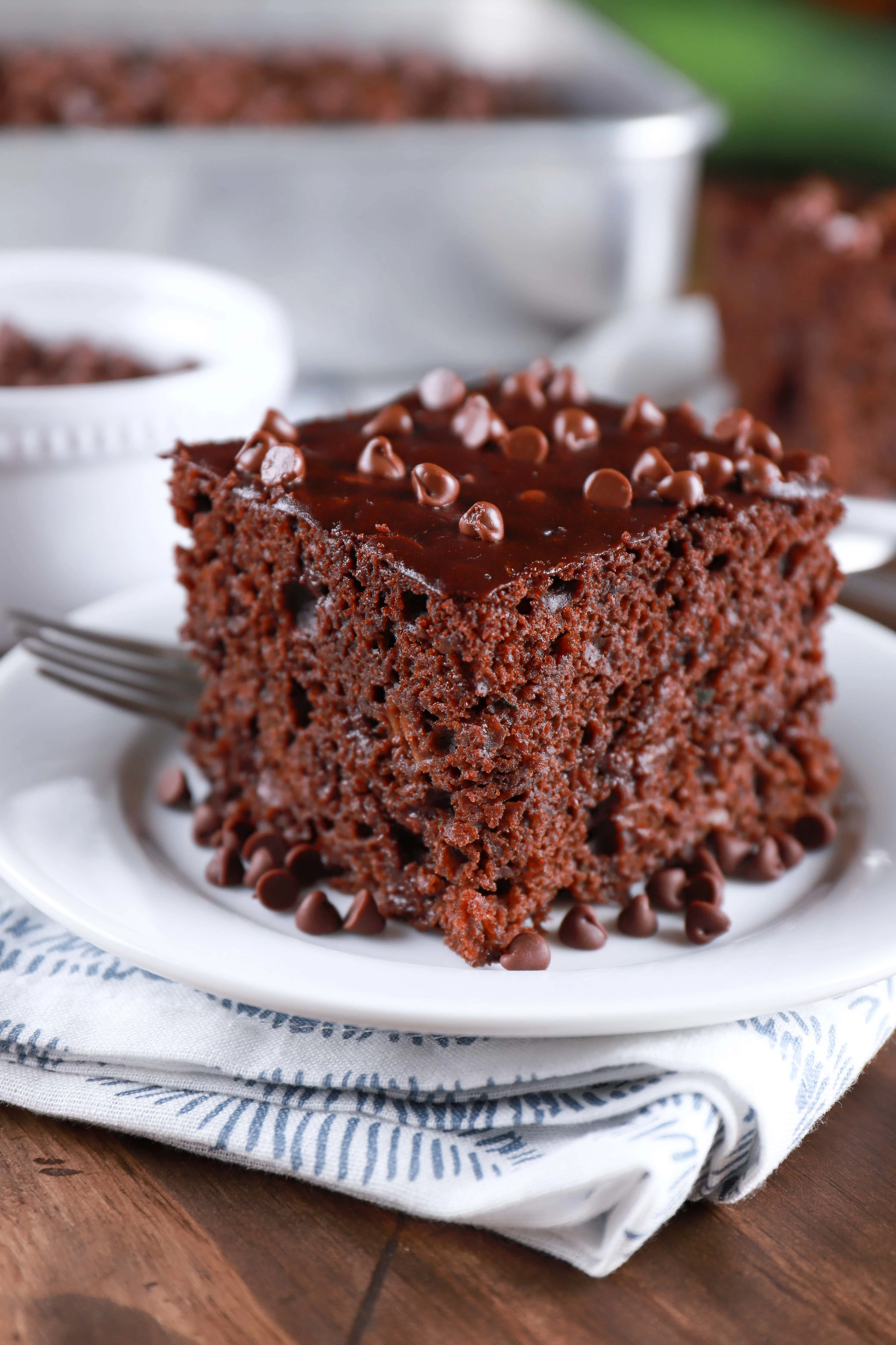 Slice of easy chocolate zucchini cake on a white plate with a pan of cake in the background. Recipe from A Kitchen Addiction