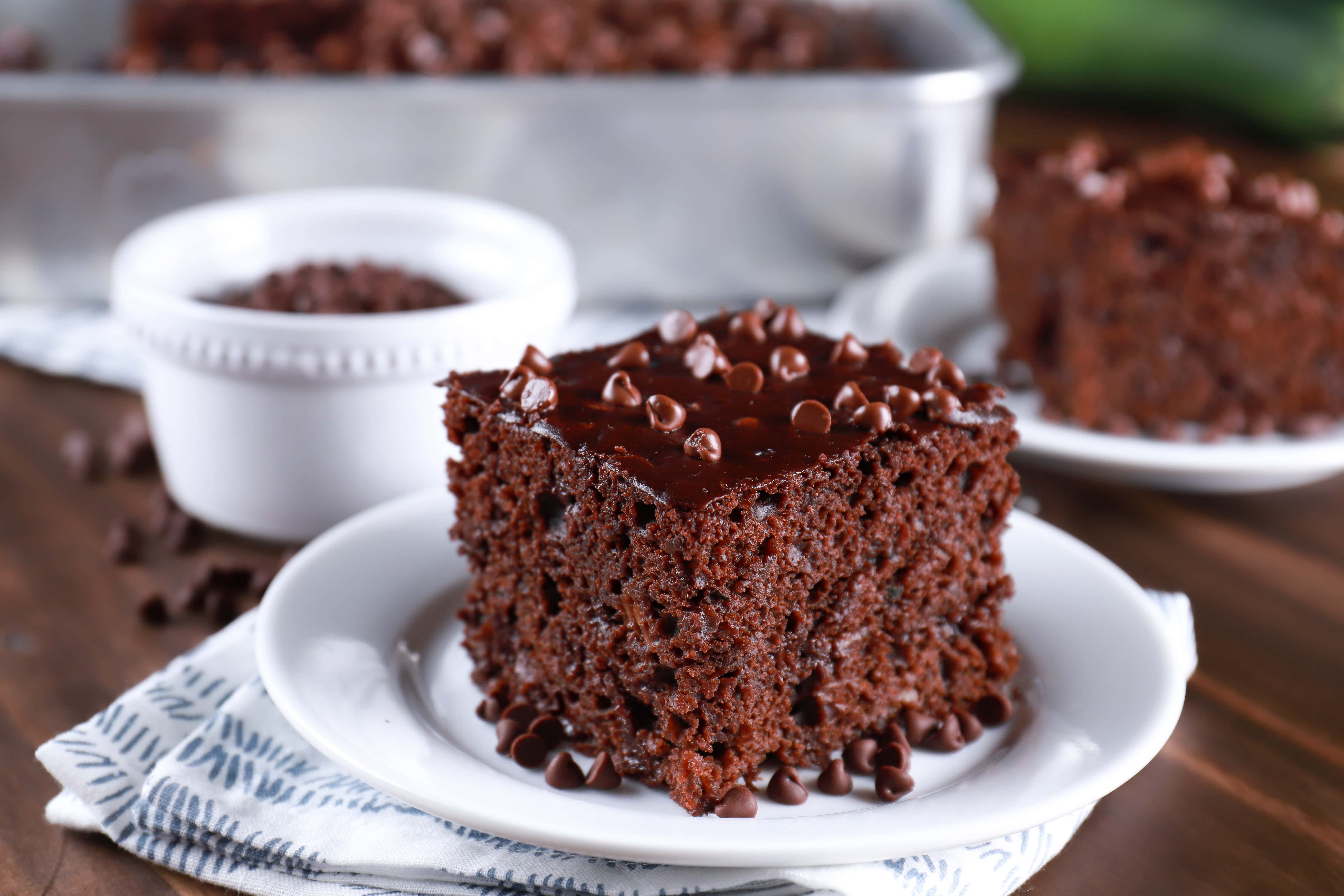 Slices of easy chocolate zucchini cake on white plates with a pan of cake in the background. Recipe for cake from A Kitchen Addiction