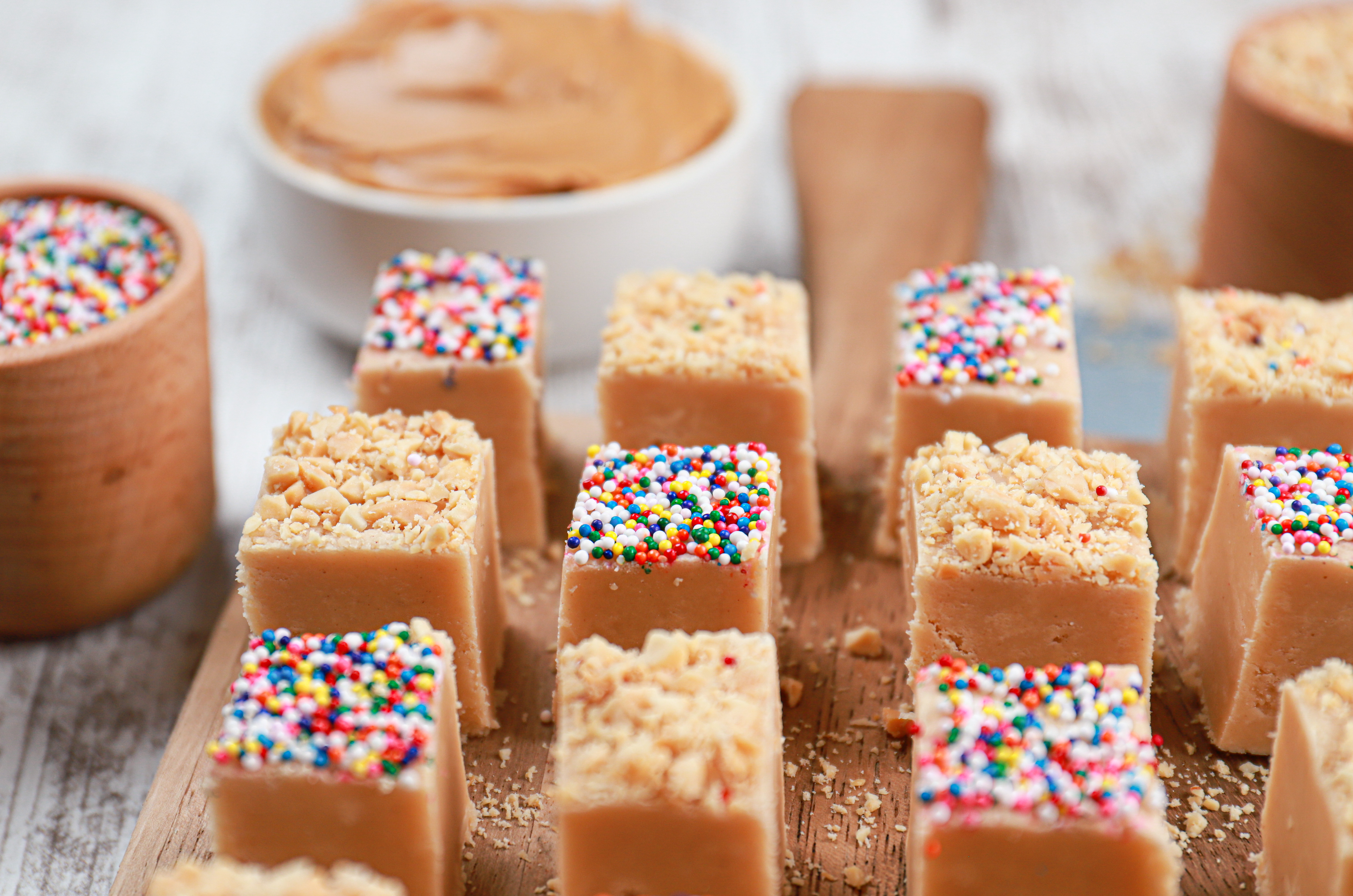 Up close view of a pieces of peanut butter fudge on a wooden cutting board with cups of sprinkles and peanut butter in the background.