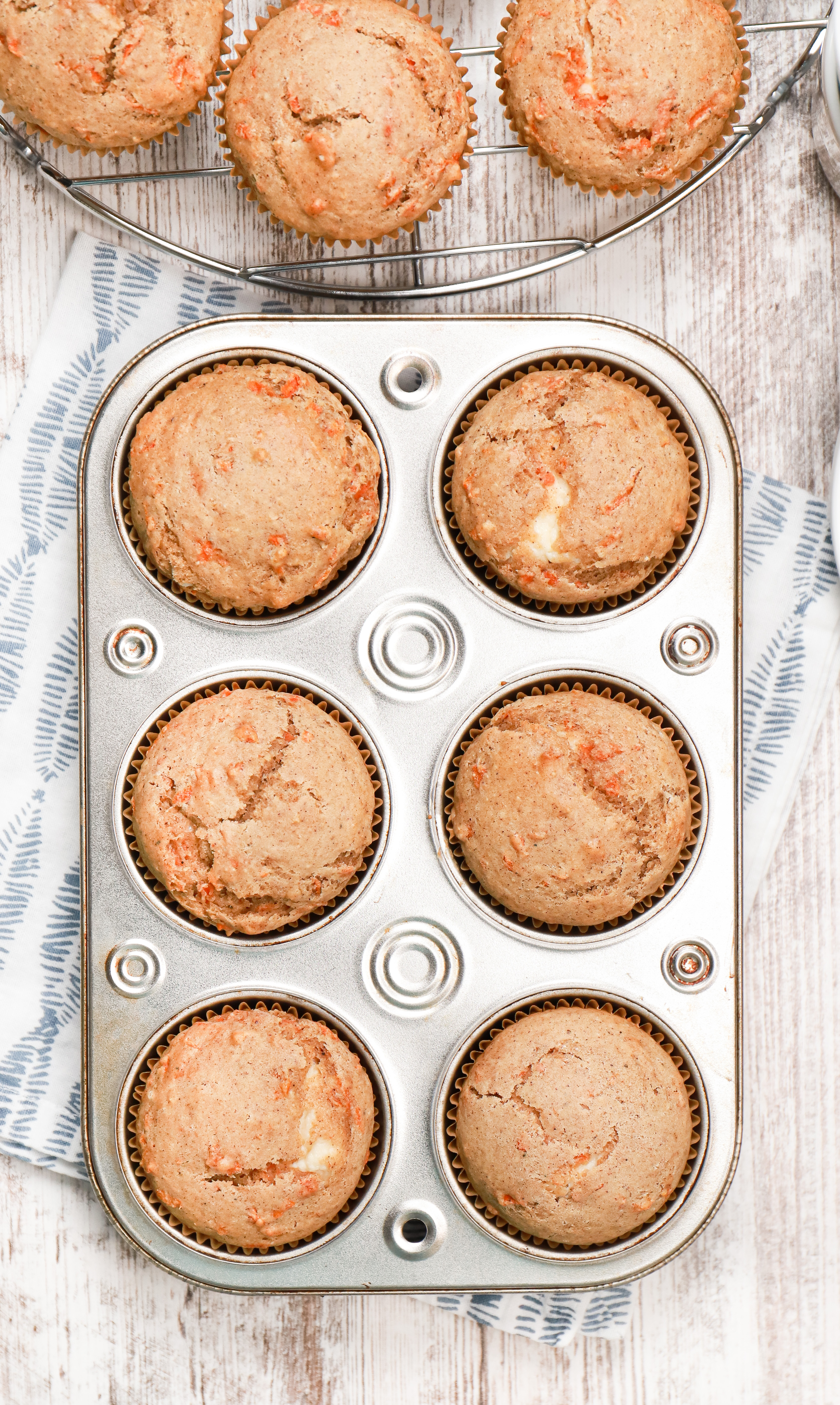 Overhead view of a batch of cream cheese filled carrot cake muffins in an aluminum muffin tin on top of a white and blue towel.