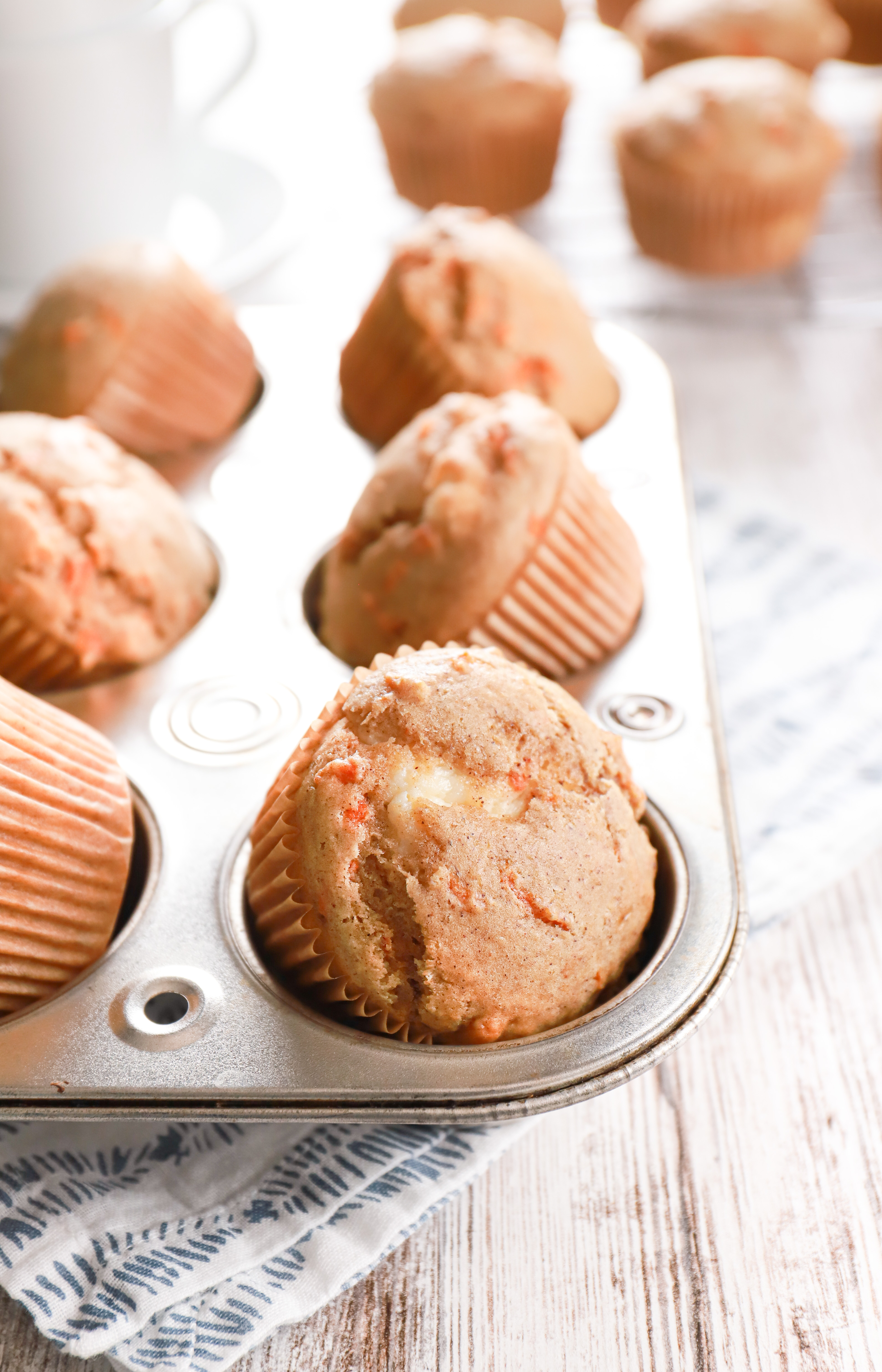 Side view of a batch of cream cheese filled carrot cake muffins in a muffin tin with a cooling rack in the background with more muffins.
