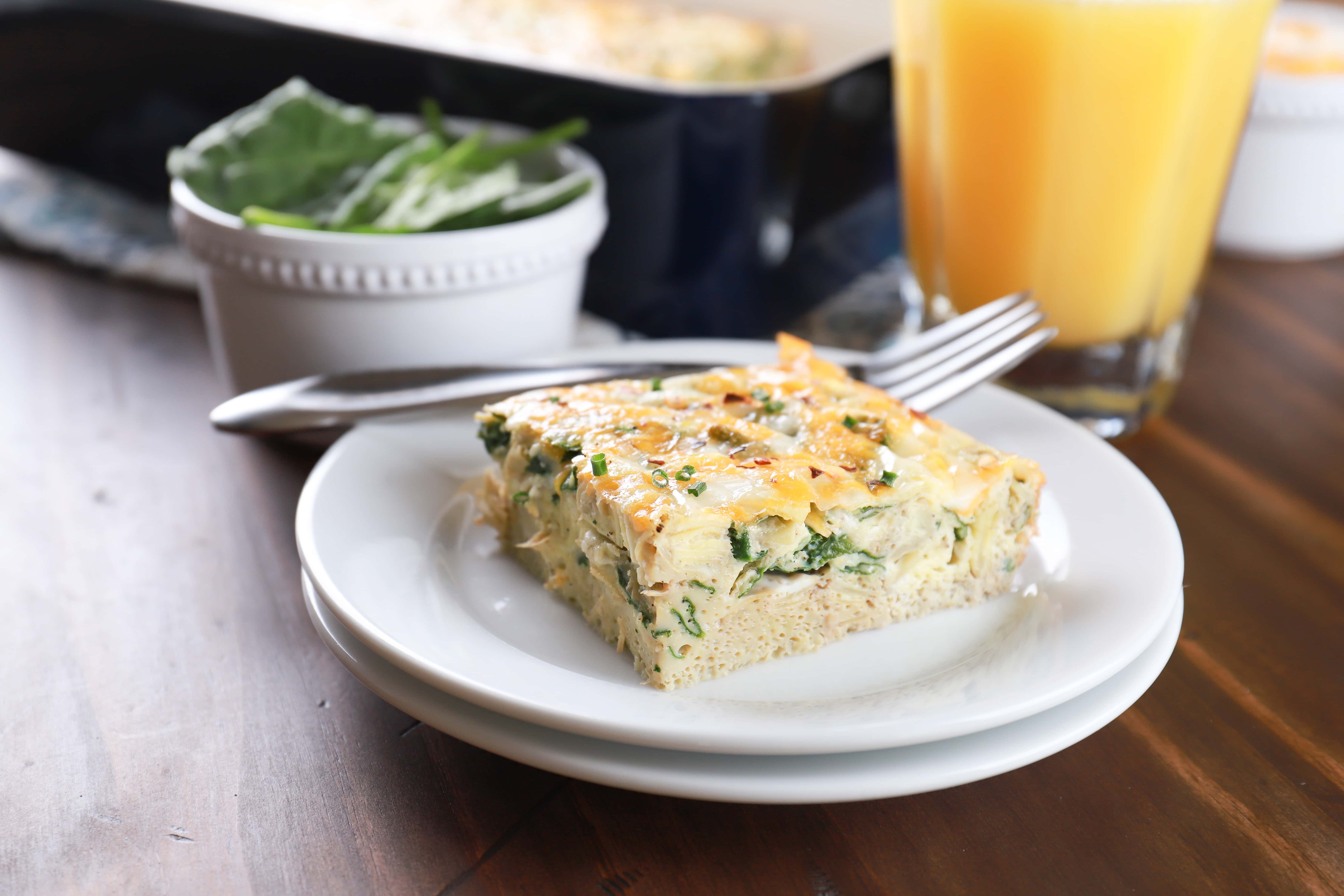 Piece of spinach artichoke egg bake on a white plate with a pan of egg bake in the background. Recipe for egg bake from A Kitchen Addiction