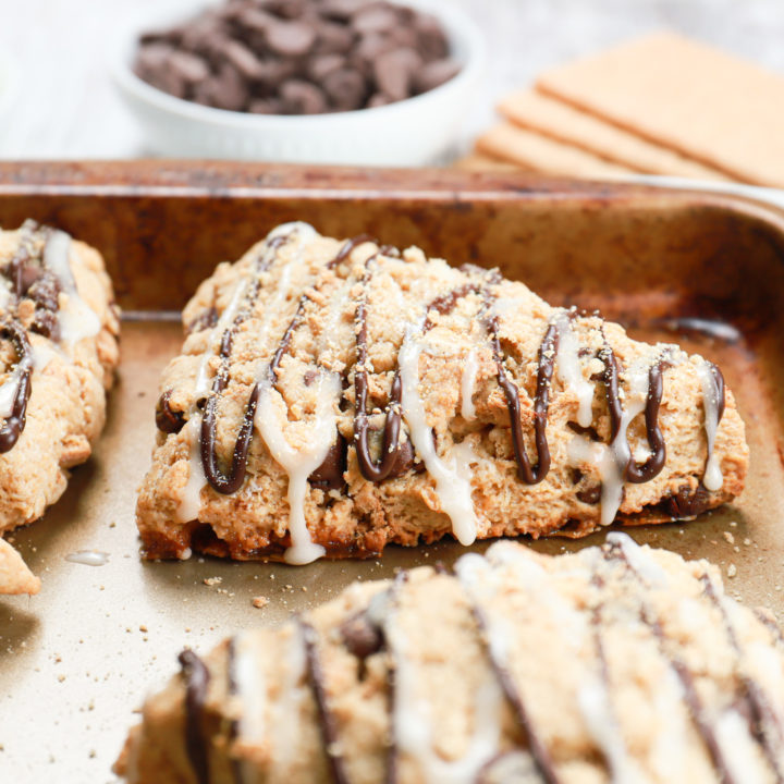 Up close side view of a s'mores scone on a baking sheet with marshmallows, chocolate chips, and graham crackers in the background.