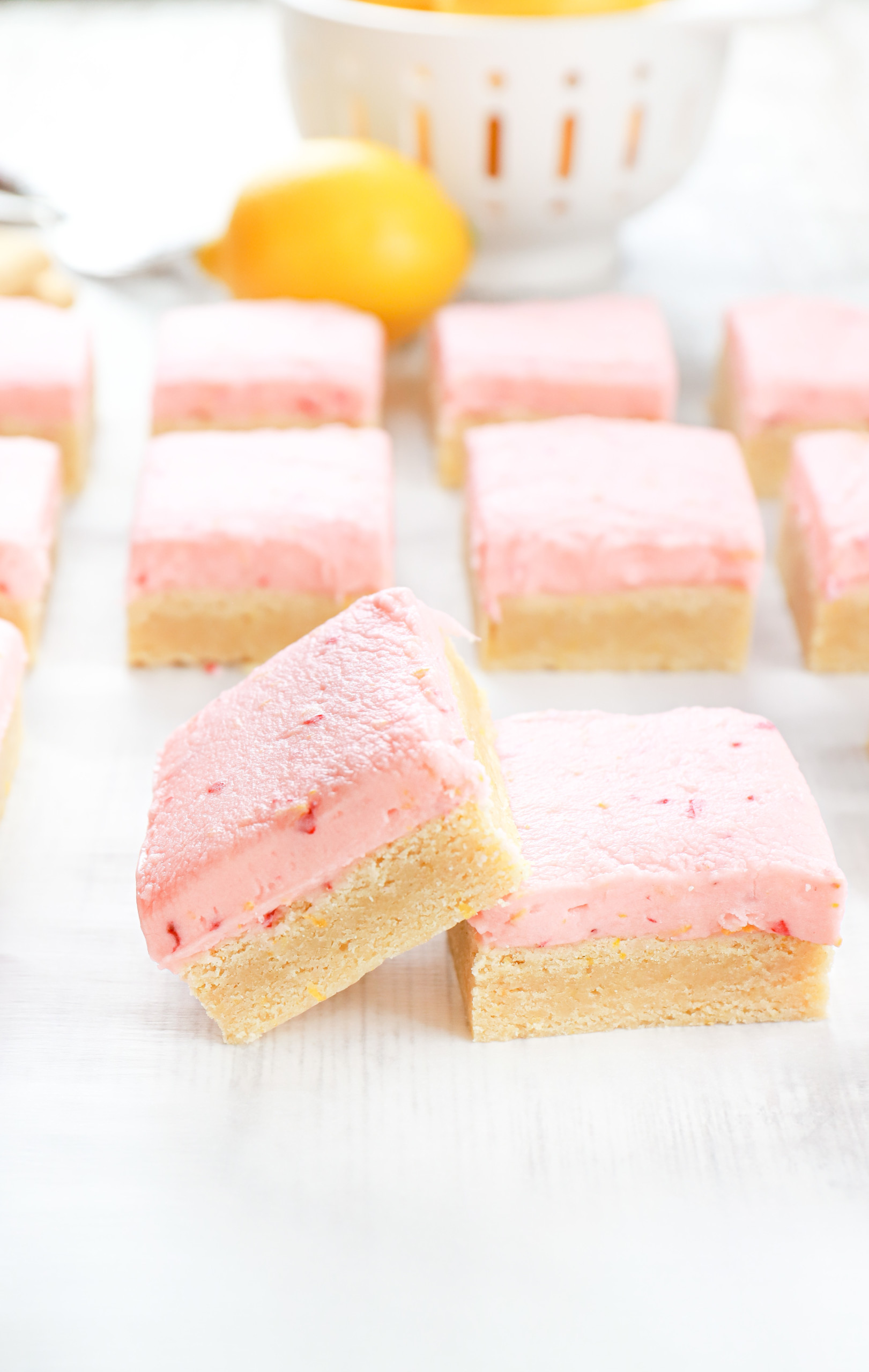 Side view of one strawberry lemon frosted sugar cookie bar propped up on another cookie bar with the remainder of the batch of bars in the background.