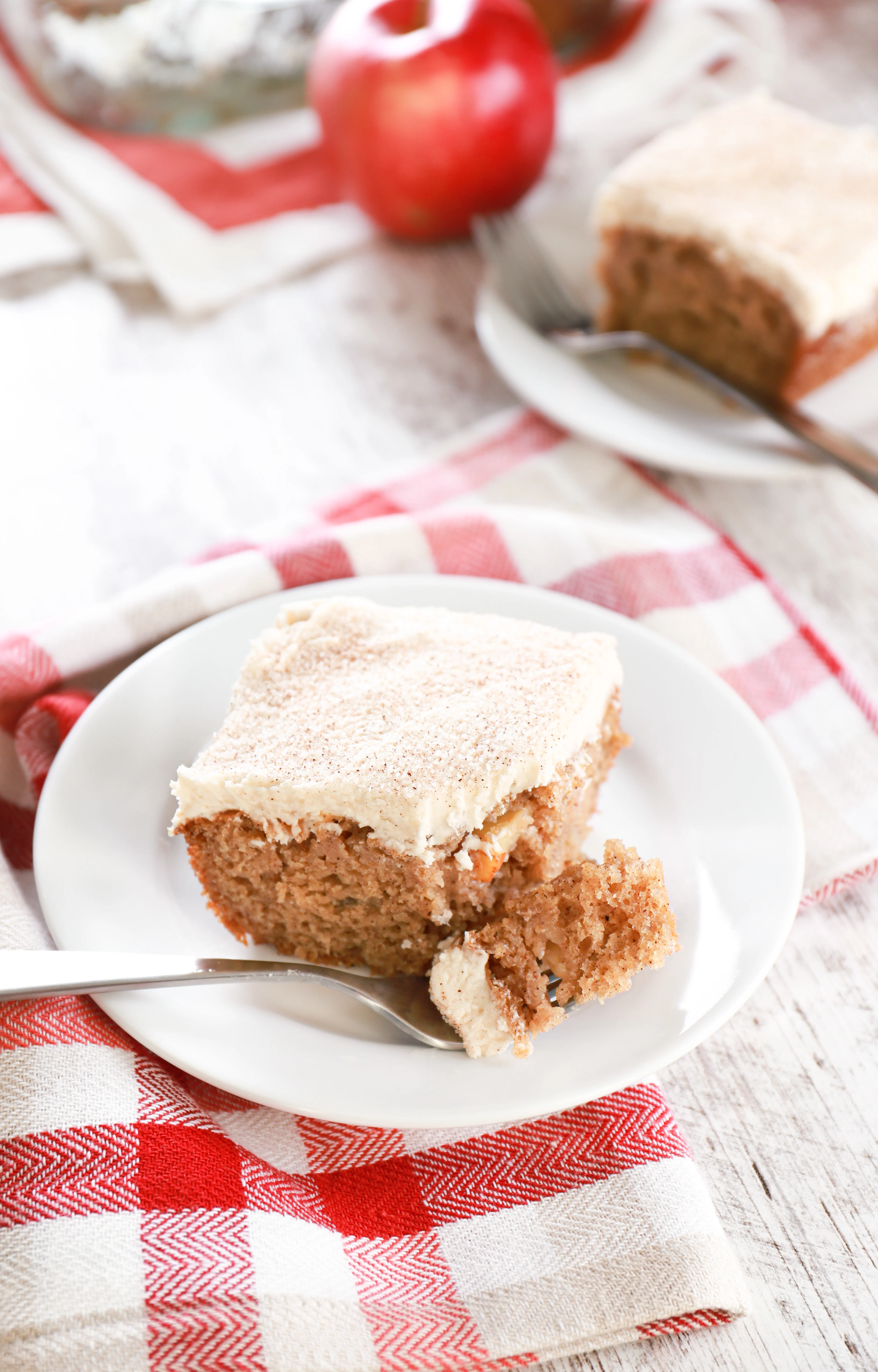 Piece of apple cake on a small white plate with a bite of cake on fork. Remaining cake in the background