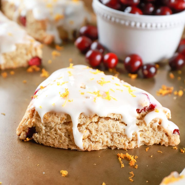 Up close view of a glazed cranberry orange scone on a baking sheet with oranges and cranberries in the background. Recipe from A Kitchen Addiction