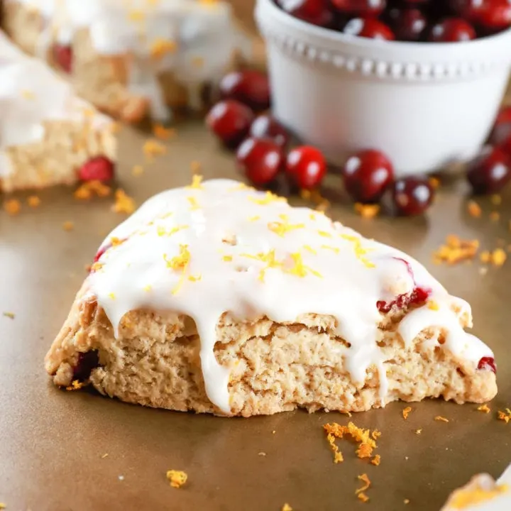 Up close view of a glazed cranberry orange scone on a baking sheet with oranges and cranberries in the background. Recipe from A Kitchen Addiction