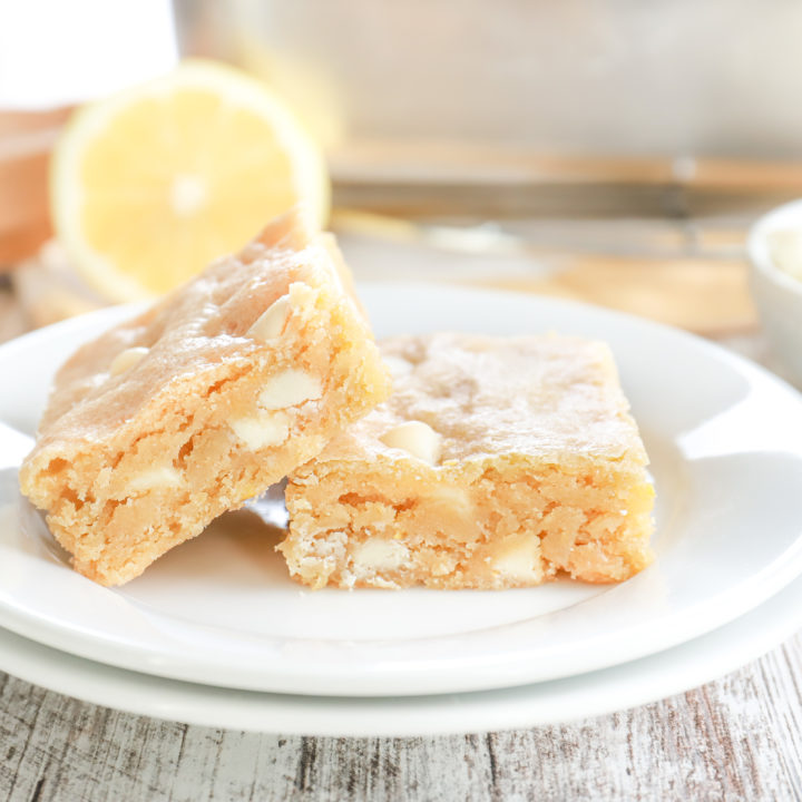 Up close side view of two white chocolate lemon blondies on a small white plate with an aluminum pan with the remaining bars in the background.