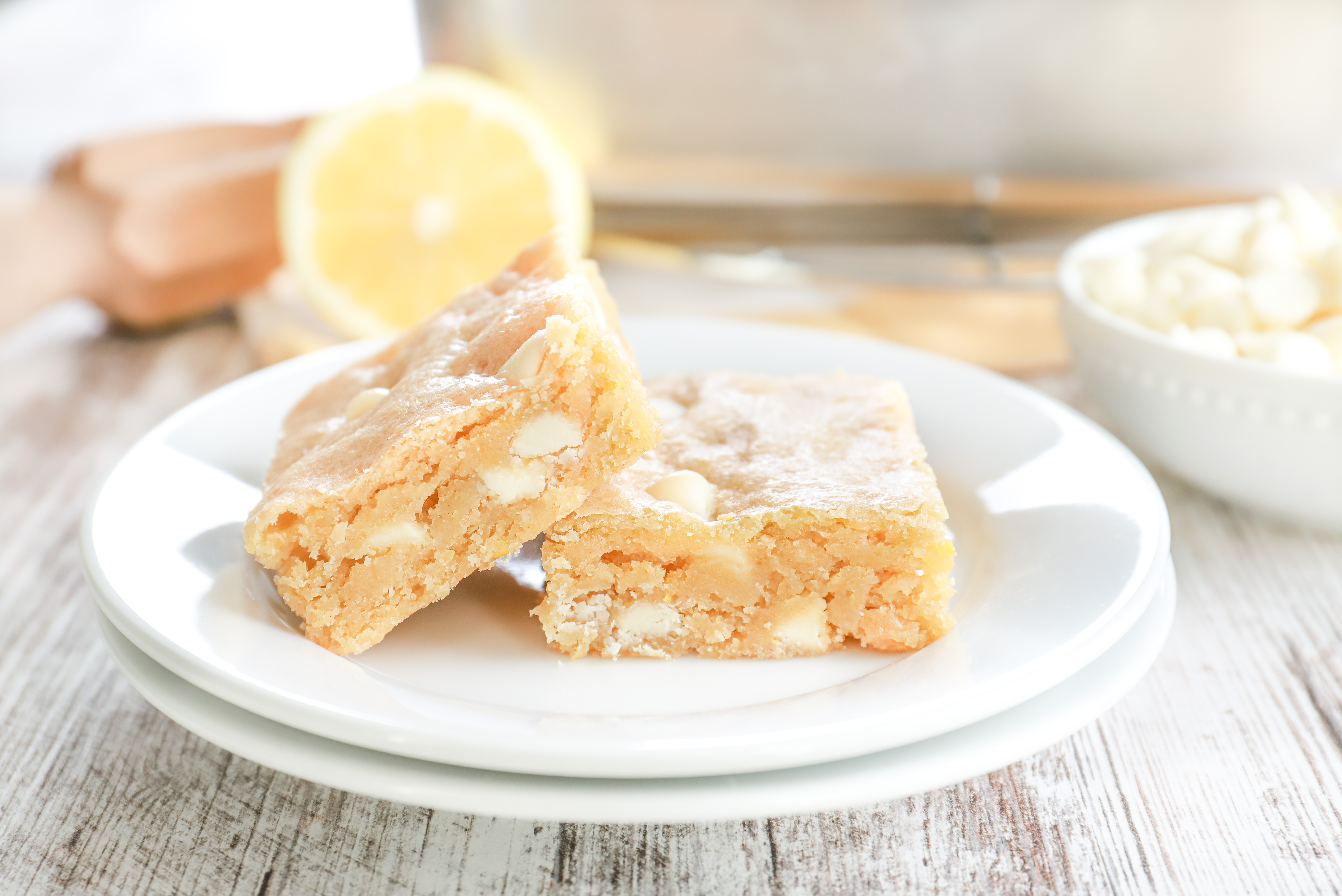 Up close side view of two white chocolate lemon blondies on a small white plate with an aluminum pan with the remaining bars in the background.