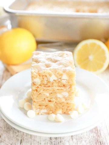 Side view of three white chocolate lemon cookie bars on a small white plate with a couple of lemons and the pan of remaining bars in the background.