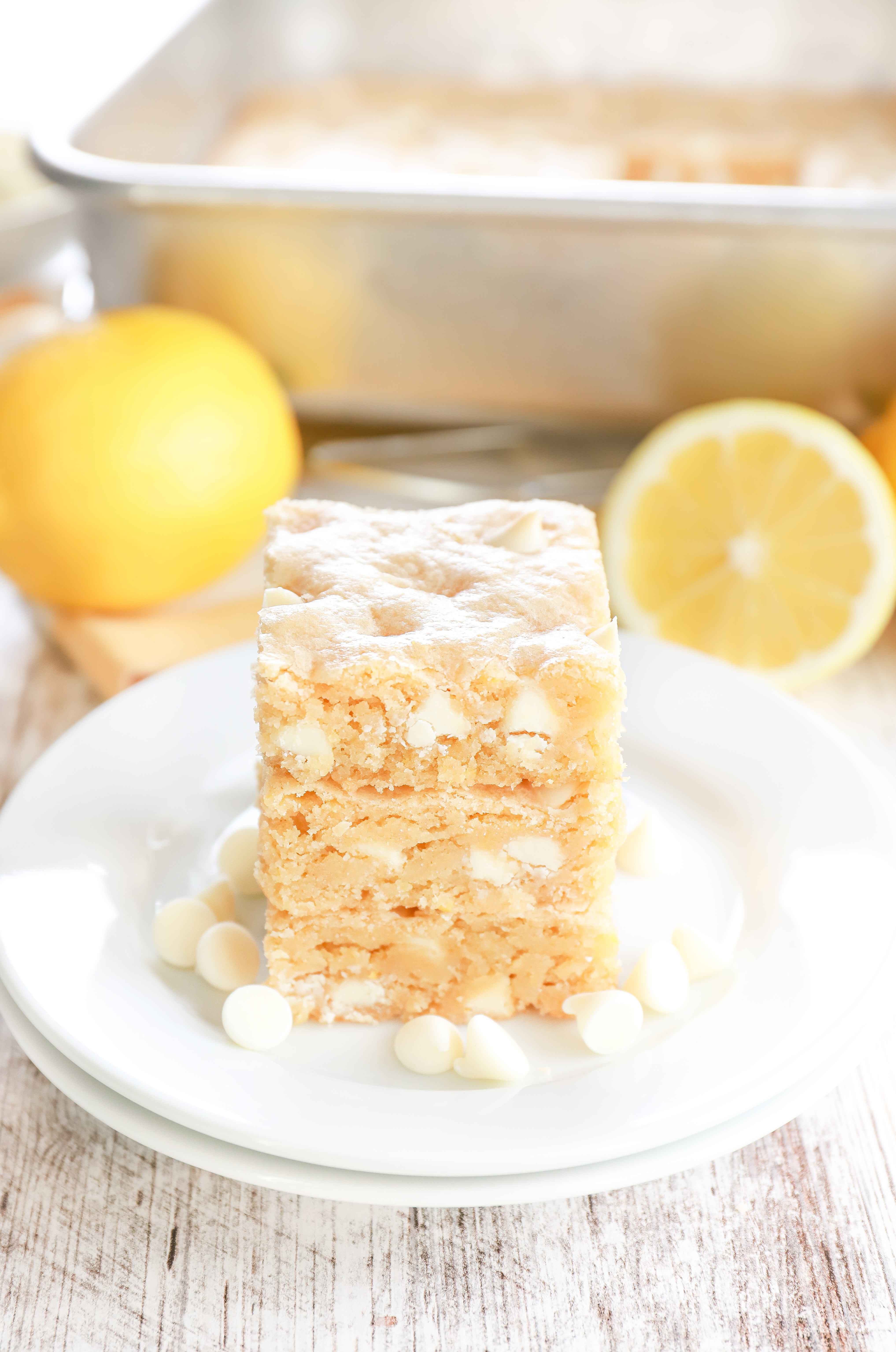 Side view of three white chocolate lemon cookie bars on a small white plate with a couple of lemons and the pan of remaining bars in the background.