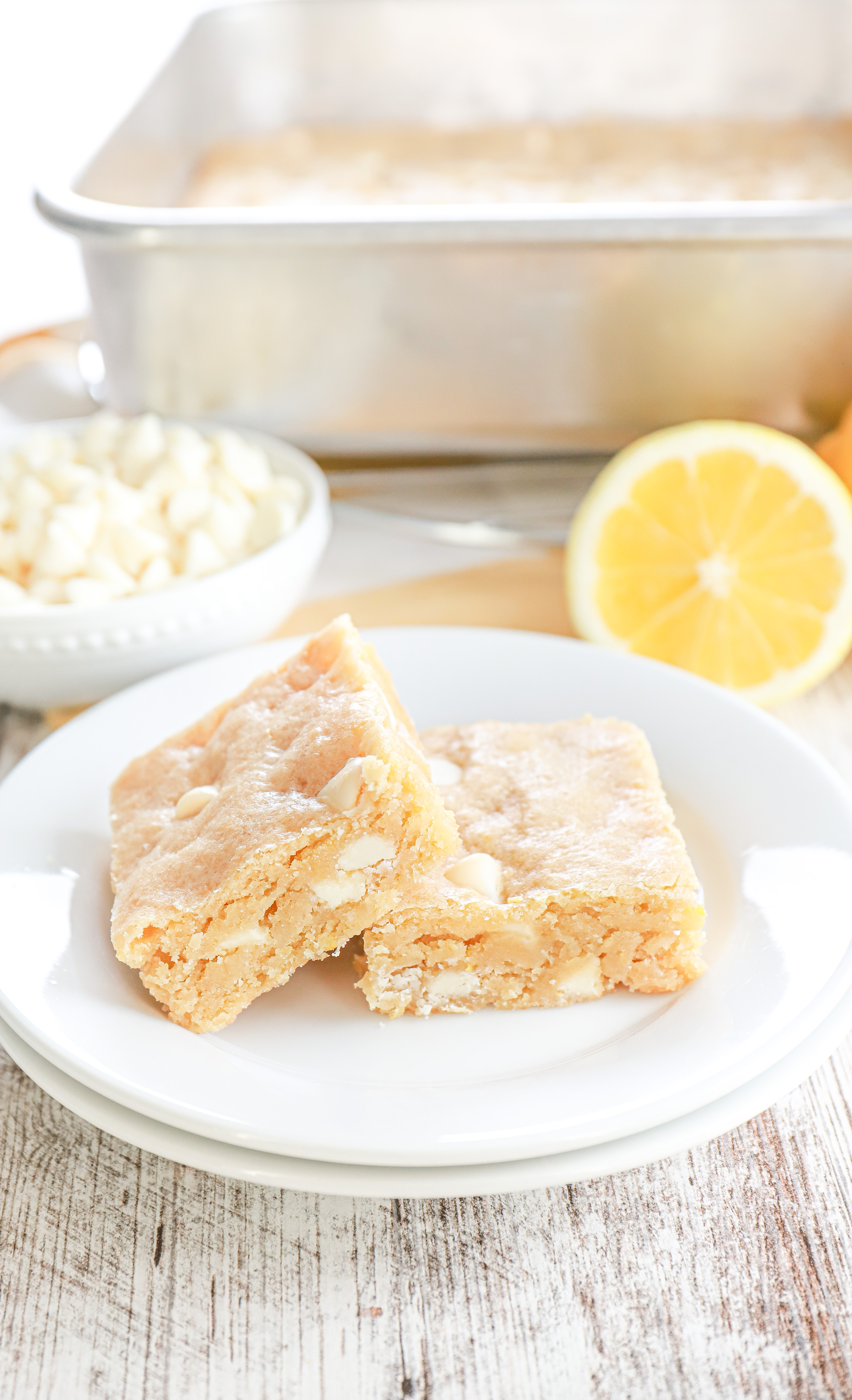 Two white chocolate lemon blondies on a small white plate with a pan of the remaining bars in the background.
