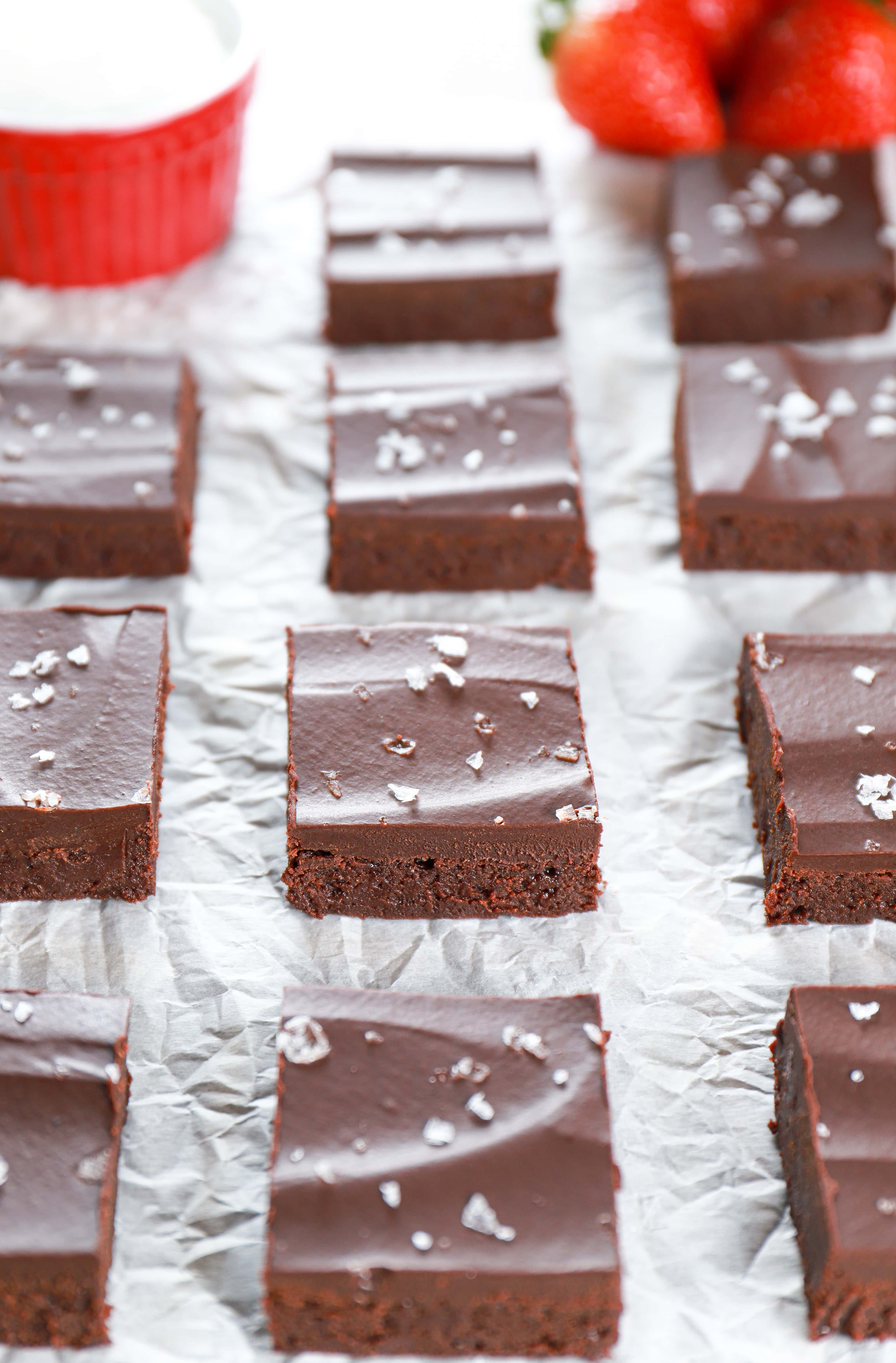 Flourless brownies with sea salt ganache lined up on a piece of parchment paper with strawberries in the background. Recipe for brownies from A Kitchen Addiction