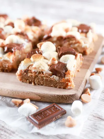 Side angle of a peanut butter smores seven layer bar on a wooden cutting board with more bars in the background. Recipe from A Kitchen Addiction