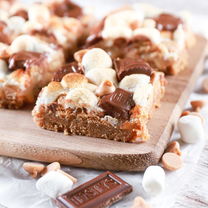 Side angle of a peanut butter smores seven layer bar on a wooden cutting board with more bars in the background. Recipe from A Kitchen Addiction