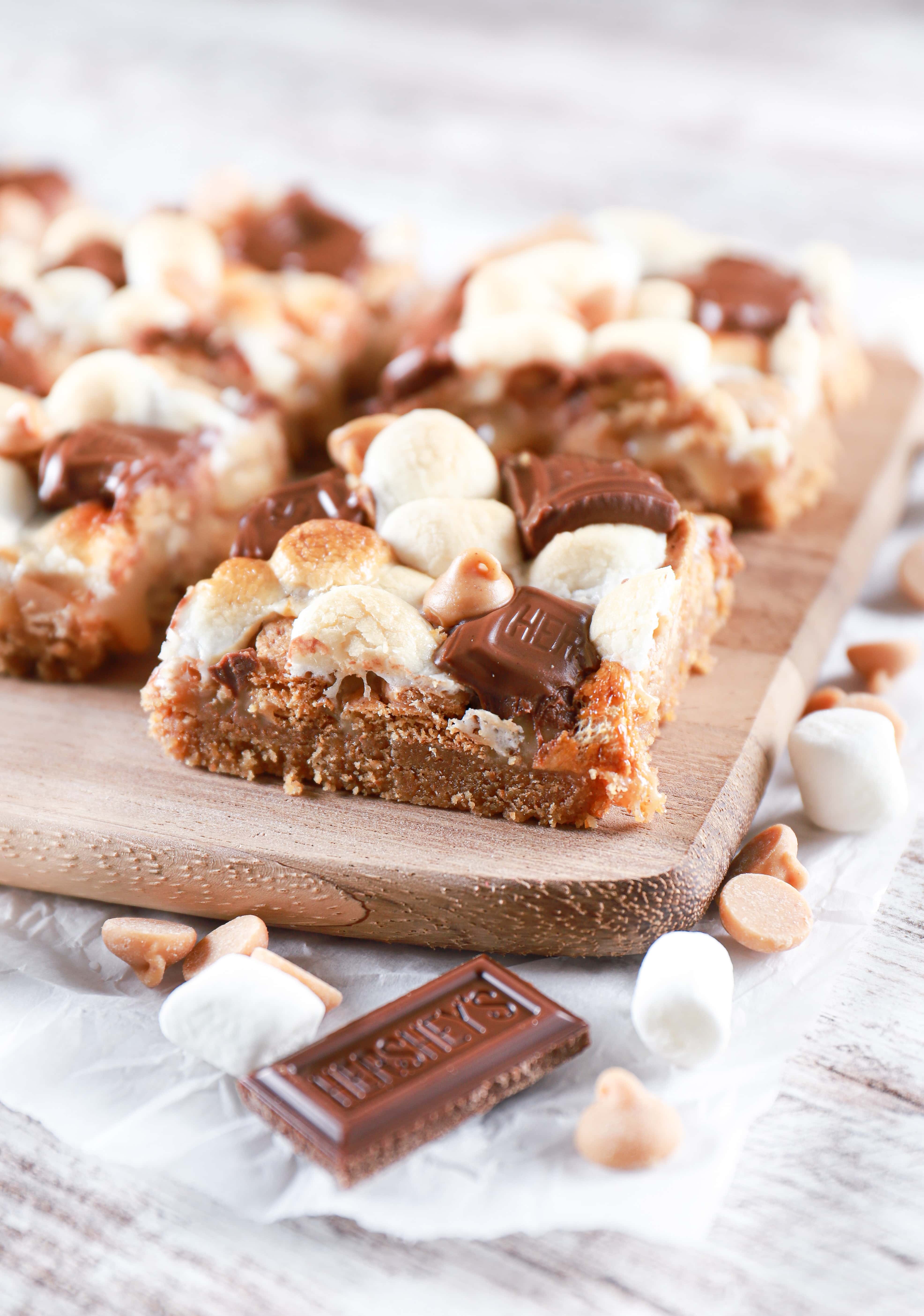 Side angle of a peanut butter smores seven layer bar on a wooden cutting board with more bars in the background. Recipe from A Kitchen Addiction