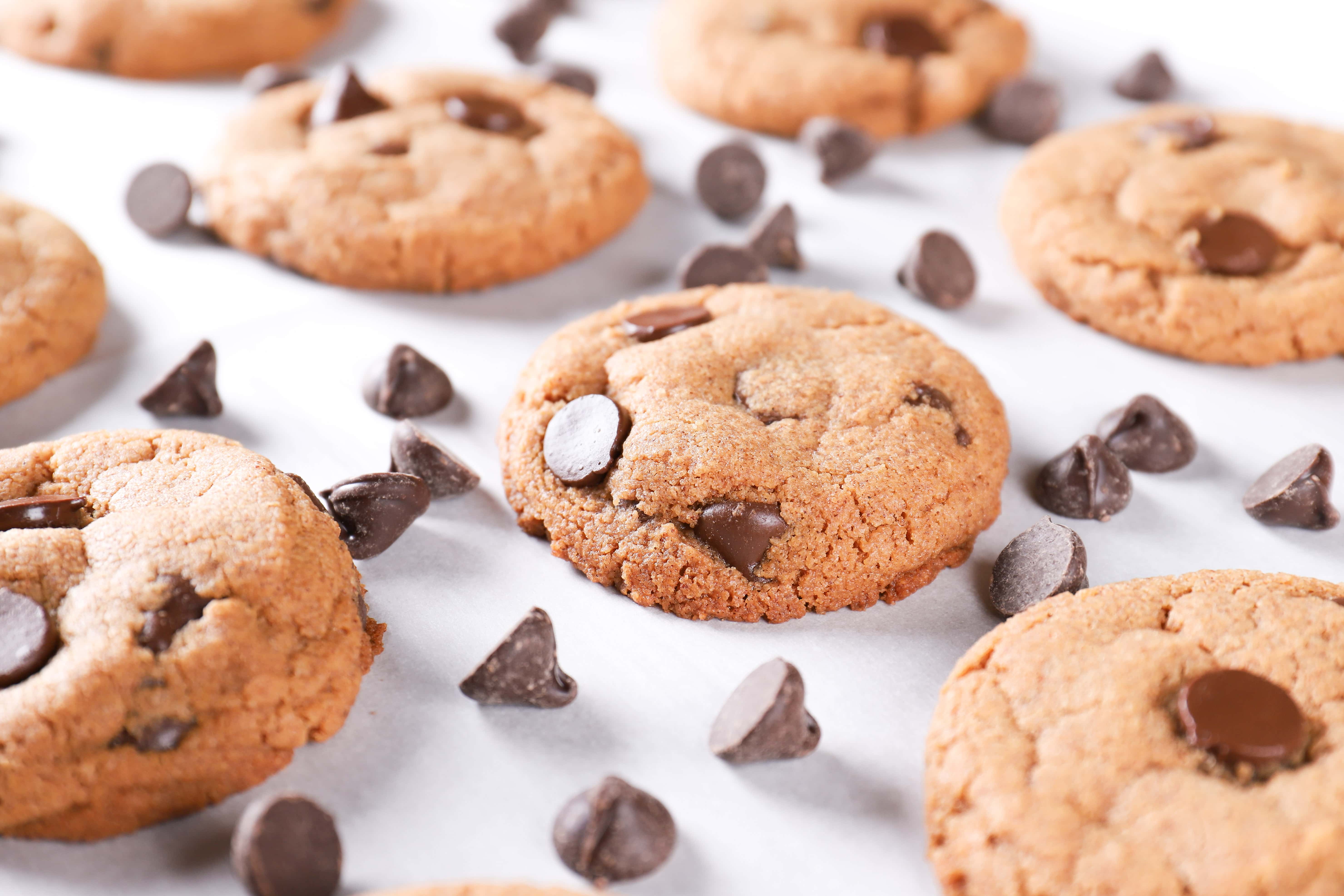 Side view of flourless almond butter chocolate chip cookies on a cookie sheet surrounded by chocolate chips. Recipe on A Kitchen Addiction