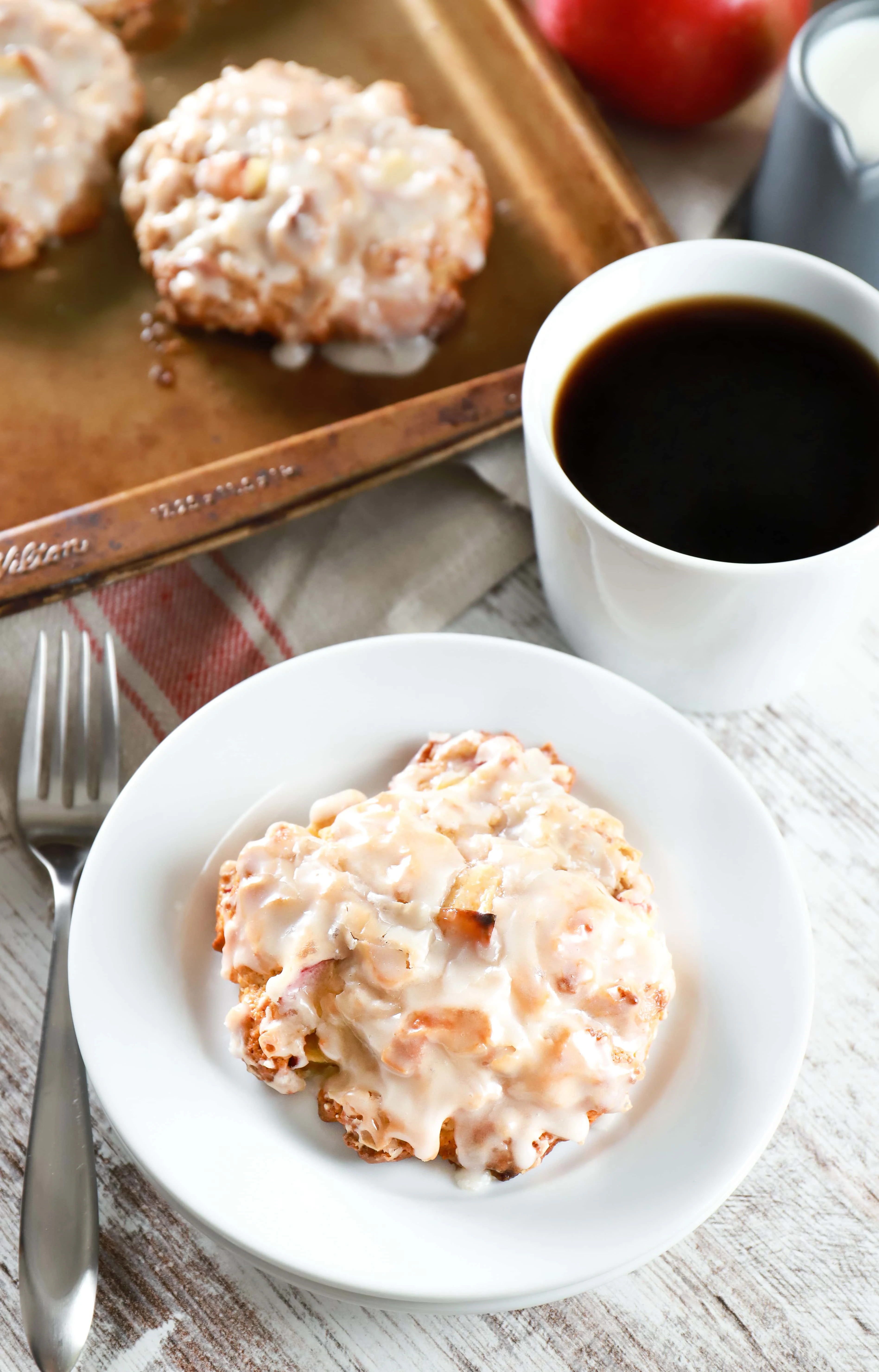 Above image of a baked apple fritter on a white plate with a baking sheet full of fritters in the background. Recipe from A Kitchen Addiction