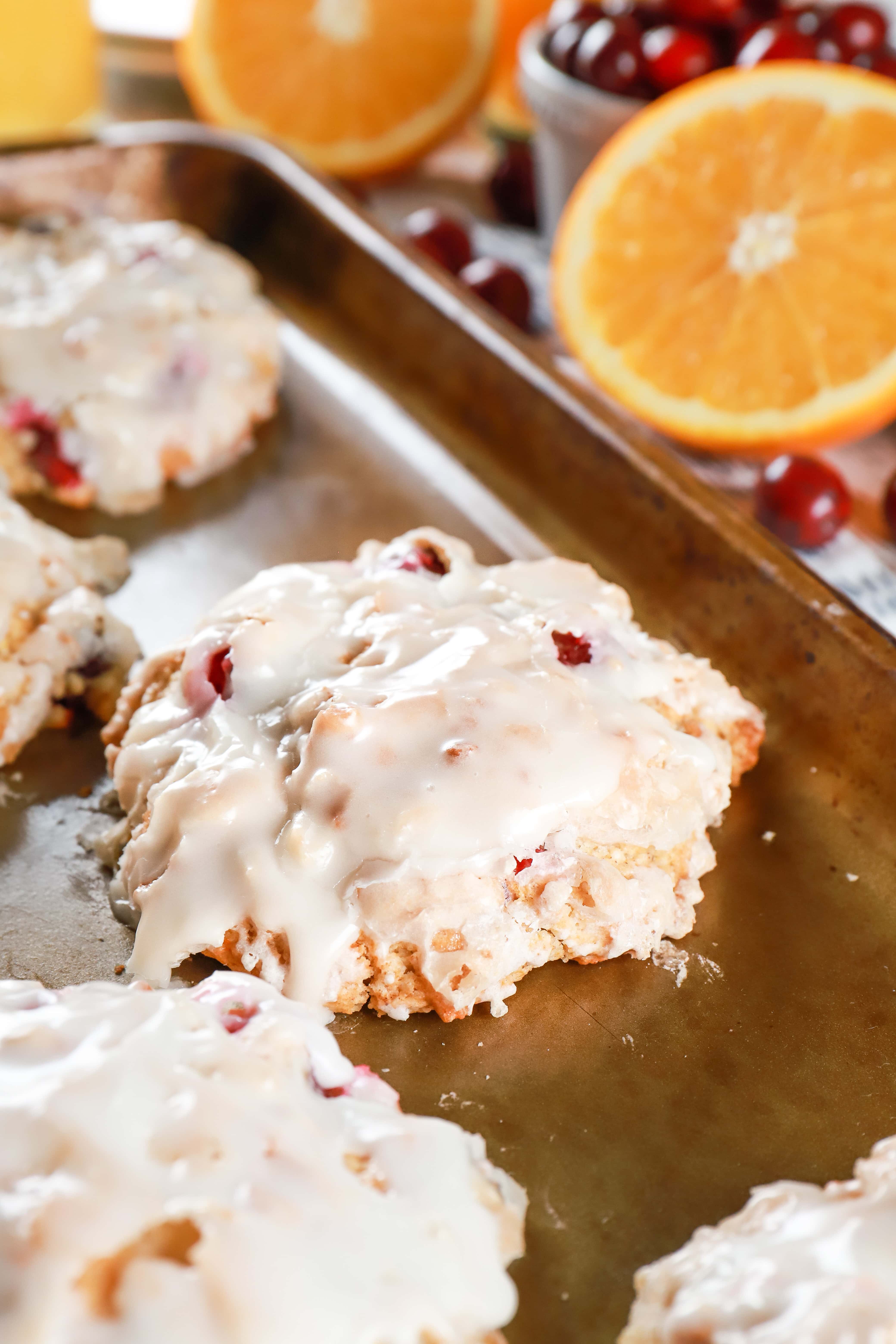 Glazed Cranberry Orange Fritters on a baking sheet with oranges and cranberries in the background. Recipe for fritters from A Kitchen Addiction