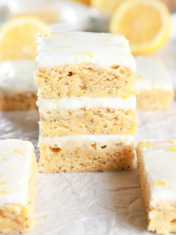 Stack of Glazed Lemon Poppy Seed Bars on parchment paper with more bars and lemons in the background. Recipe from A Kitchen Addiction