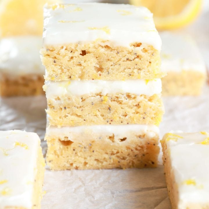 Stack of Glazed Lemon Poppy Seed Bars on parchment paper with more bars and lemons in the background. Recipe from A Kitchen Addiction