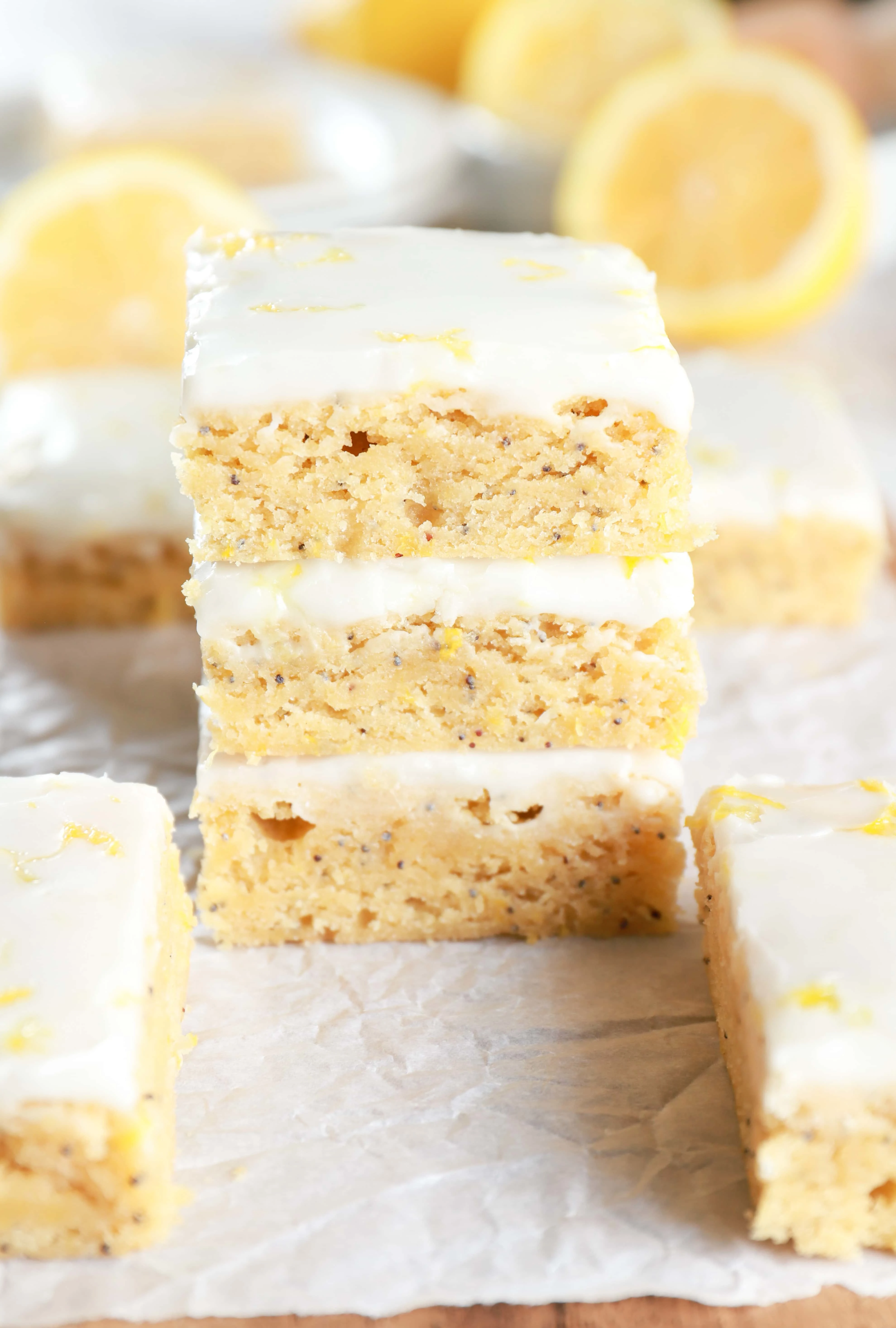 Stack of Glazed Lemon Poppy Seed Bars on parchment paper with more bars and lemons in the background. Recipe from A Kitchen Addiction