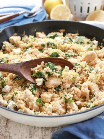 brown wooden spoon with a scoop of lemon feta chicken quinoa skillet in a white cast iron skillet on top of a blue kitchen towel