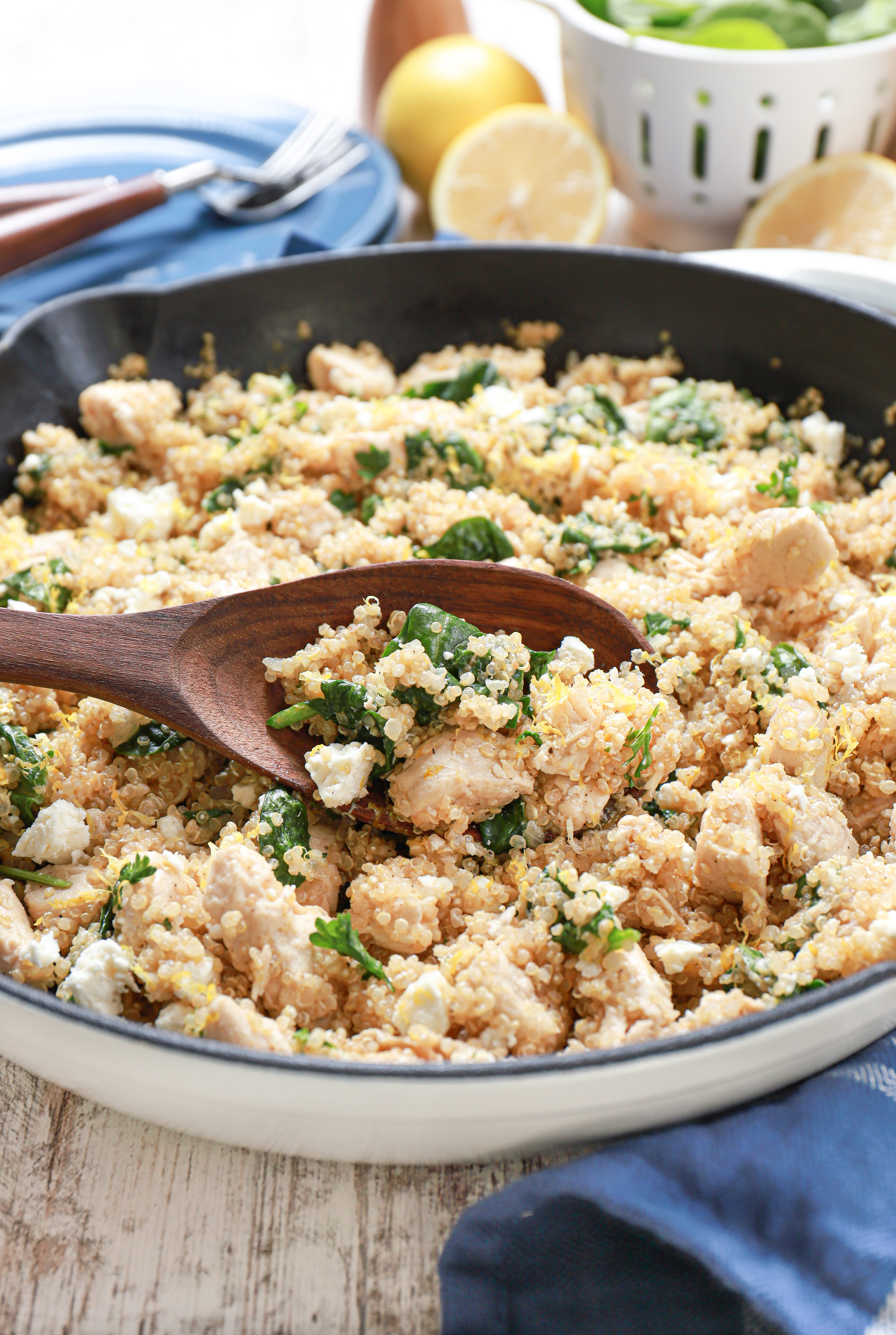 brown wooden spoon with a scoop of lemon feta chicken quinoa skillet in a white cast iron skillet on top of a blue kitchen towel