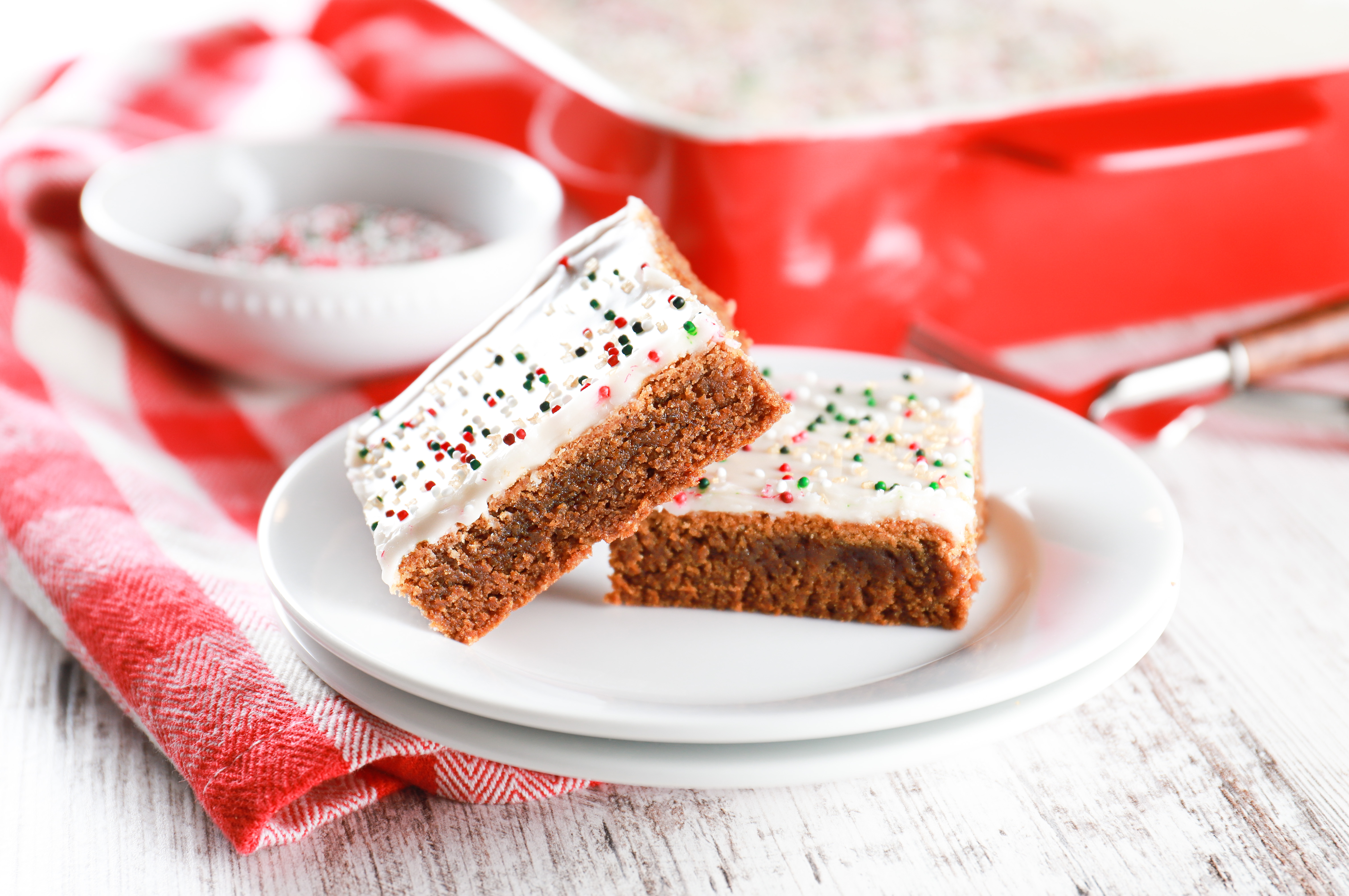 Two soft baked gingerbread bars on a small white plate with a baking dish with the remainder of the bars in the background.