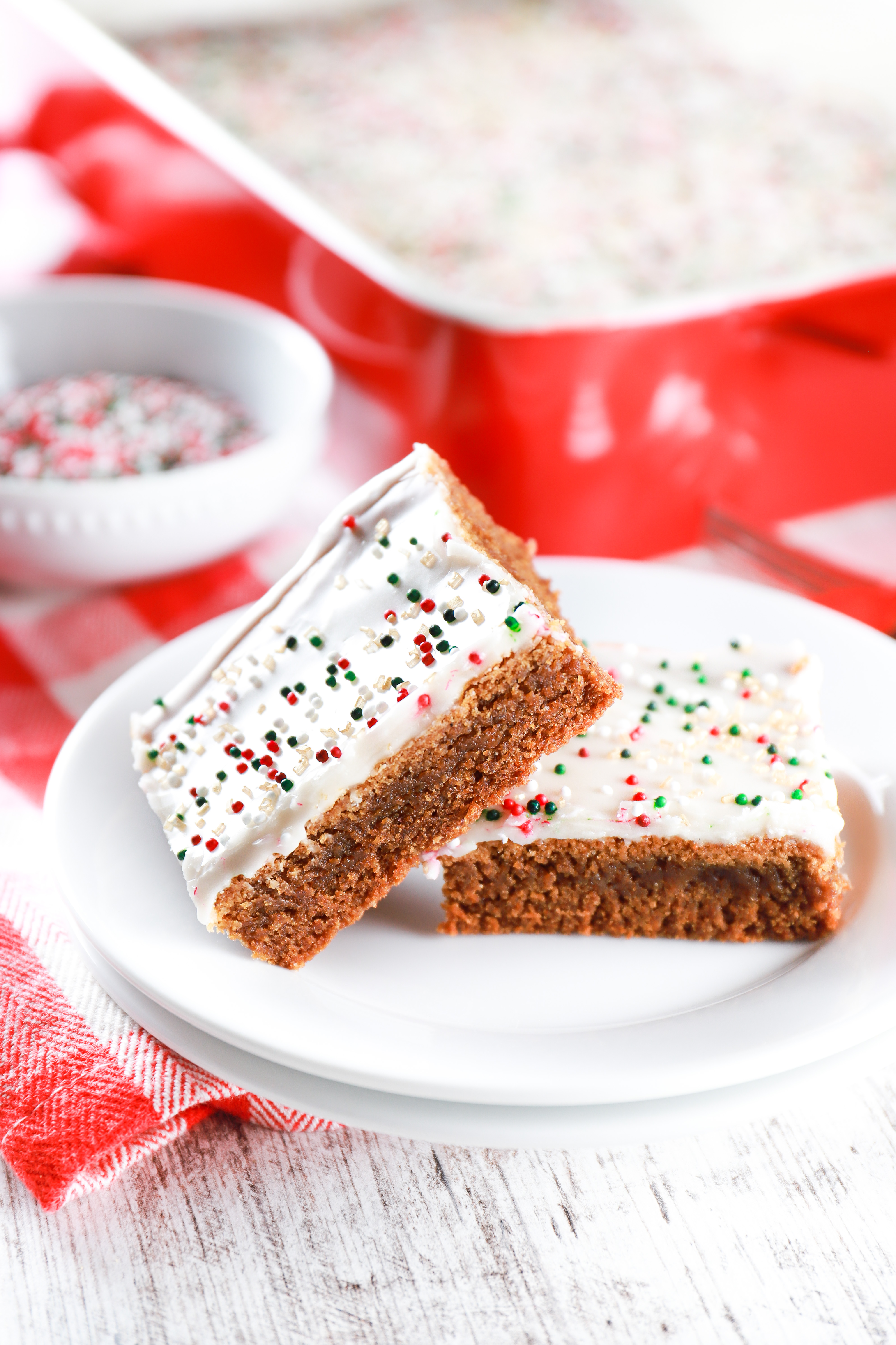 Two glazed gingerbread bars on a small white plate with a red baking dish in the background.