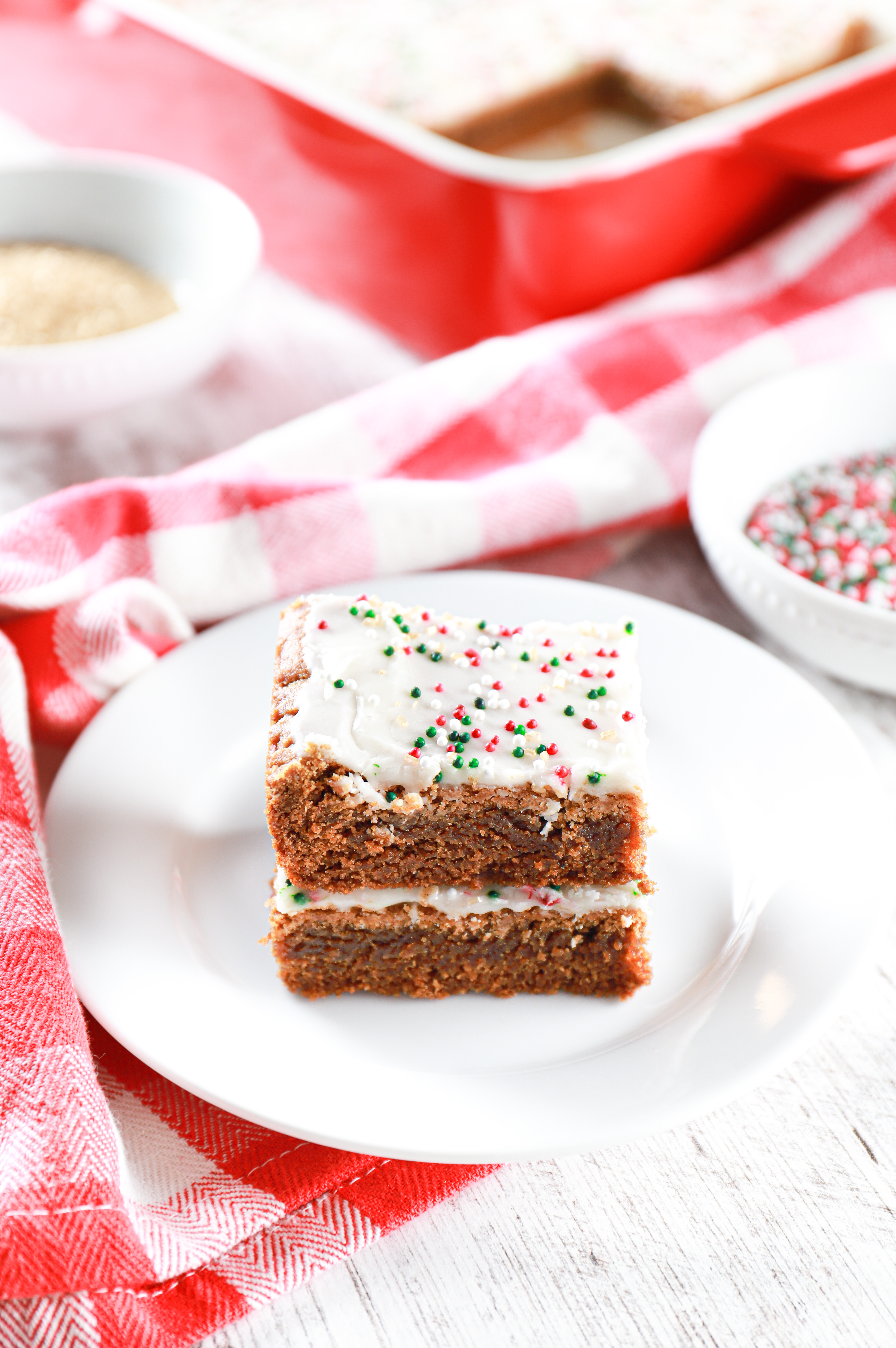 Stack of two glazed gingerbread cookie bars on a small white plate with a small bowl of red and green and white sprinkles in the background.