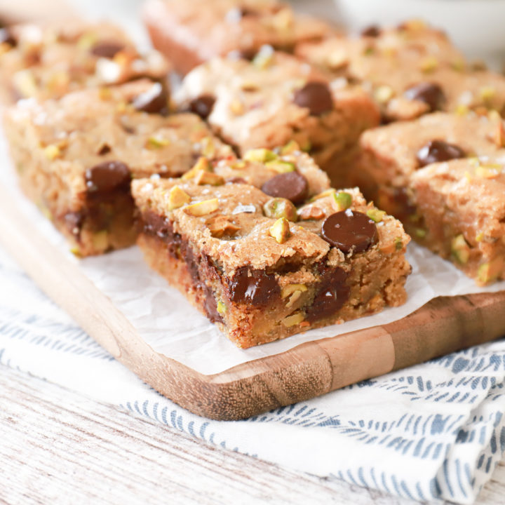 Side view of a salted dark chocolate pistachio cookie bar on a parchment paper covered wooden cutting board with more cookie bars in the background.