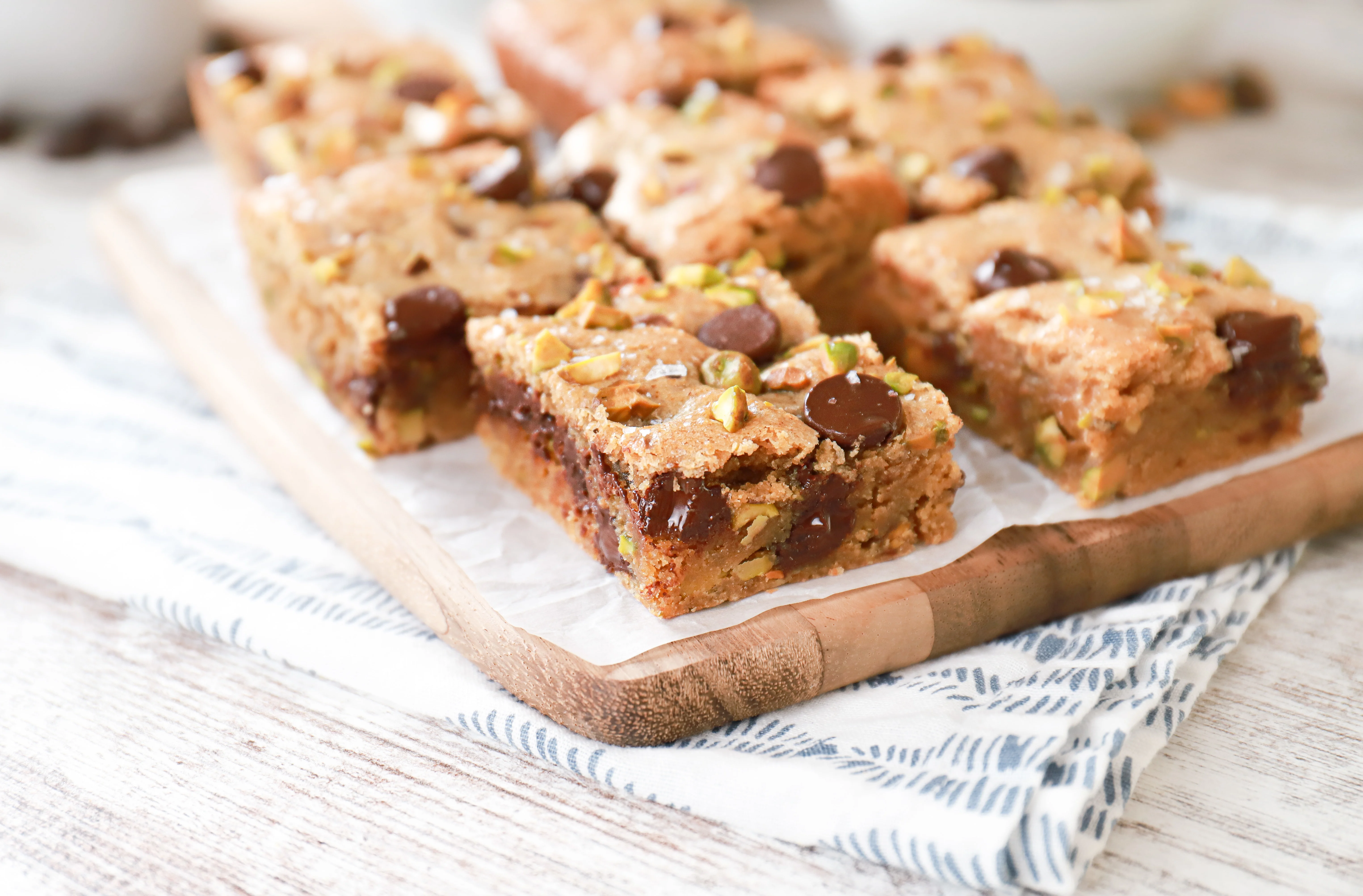 Side view of a salted dark chocolate pistachio cookie bar on a parchment paper covered wooden cutting board with more cookie bars in the background.