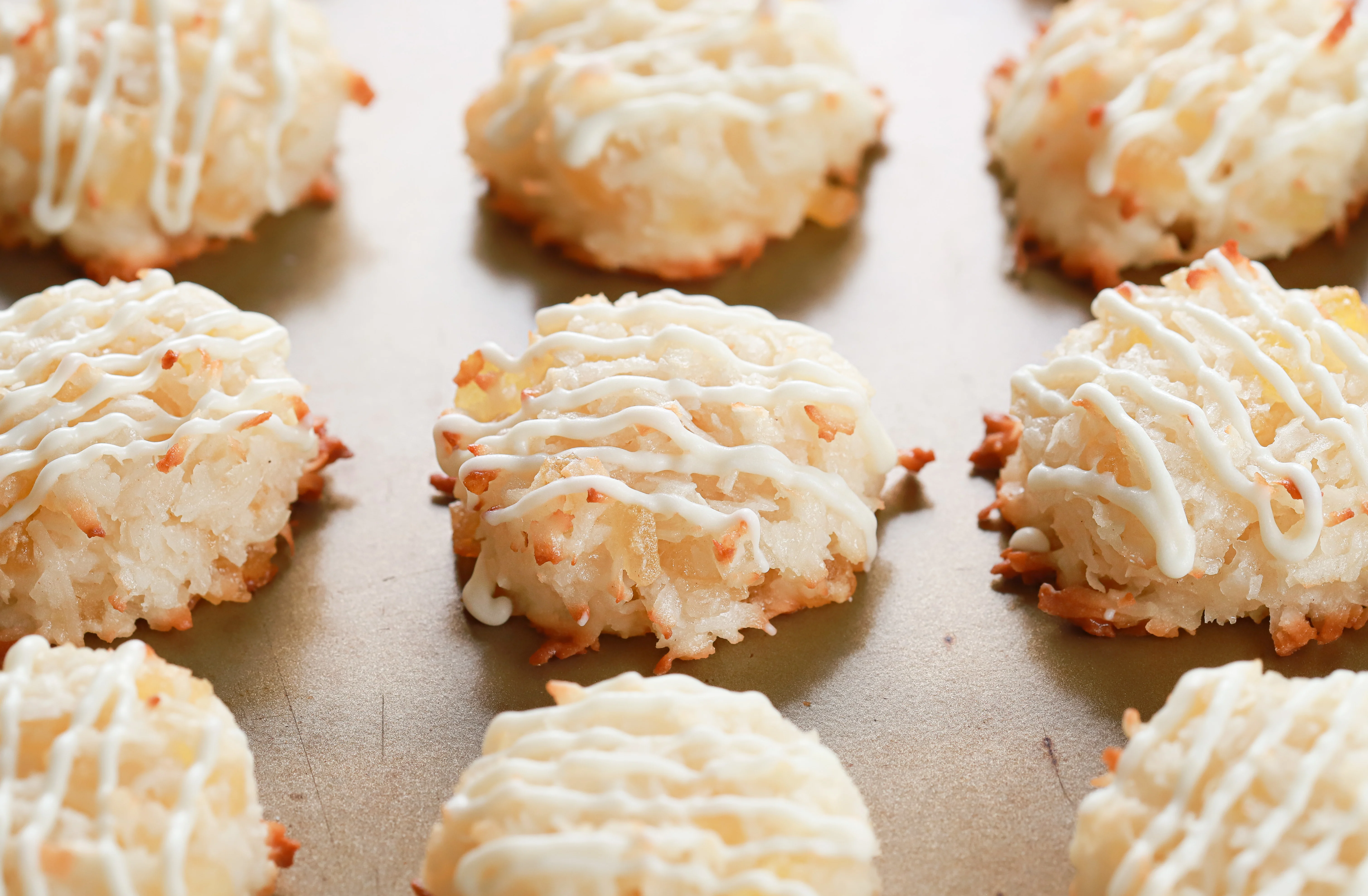 Up close side view of a white chocolate drizzled pineapple coconut macaroon on a baking sheet surrounded by other macaroons.