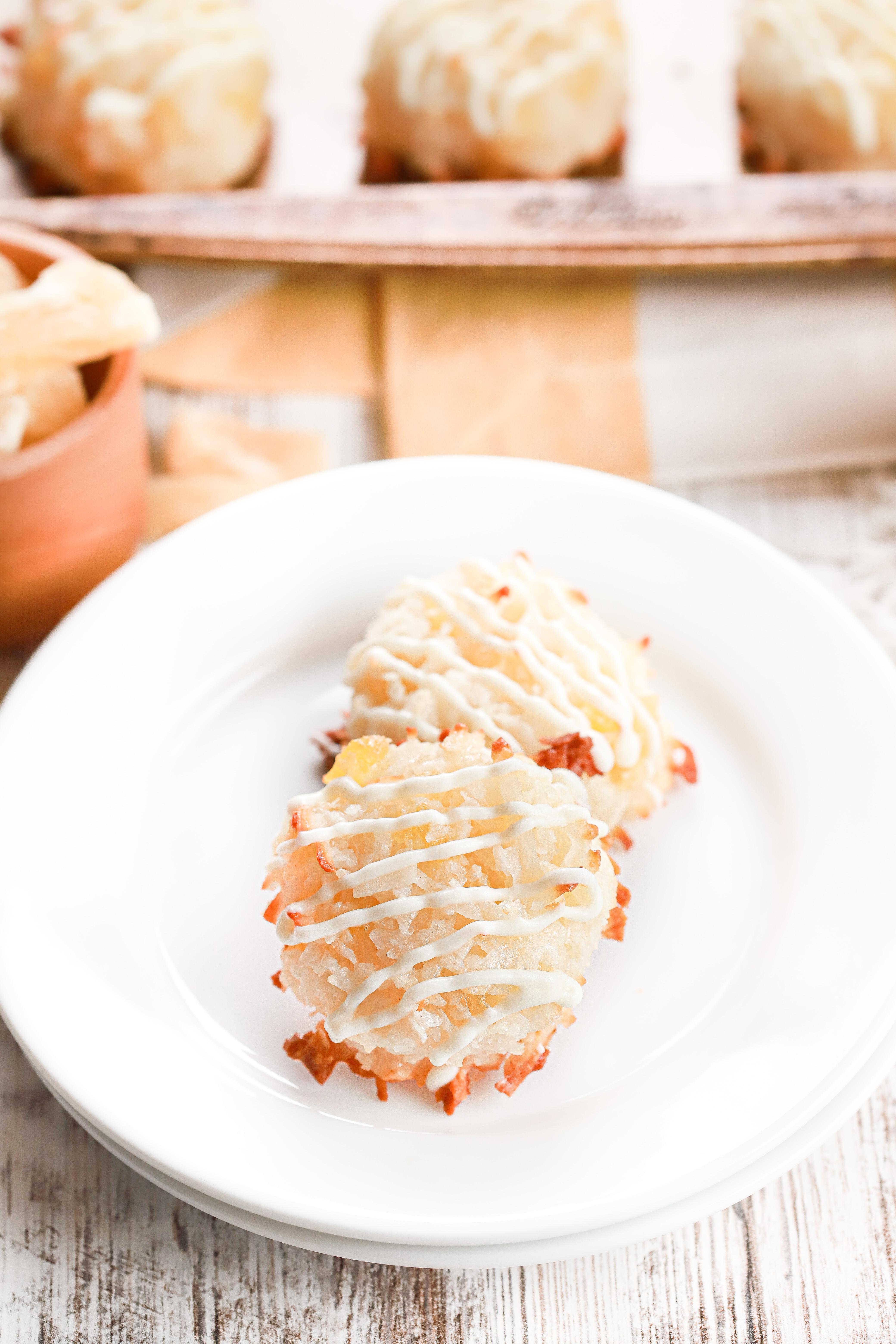 Two pineapple coconut macaroons on a small white plate with a baking sheet full of macaroons in the background.