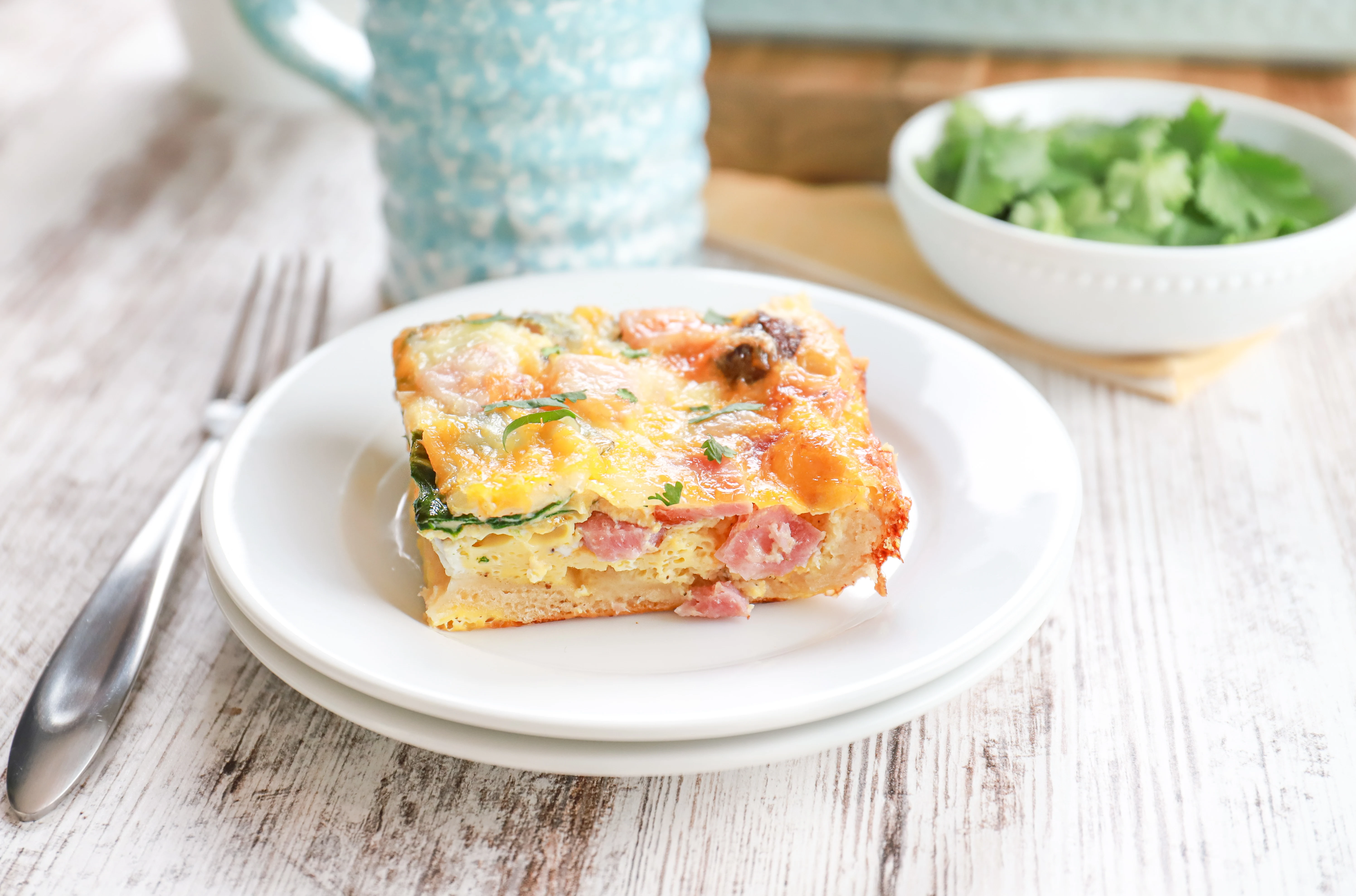 Side view of a piece of cheesy ham crescent roll breakfast bake on a small white plate with a small white bowl of parsley in the background.