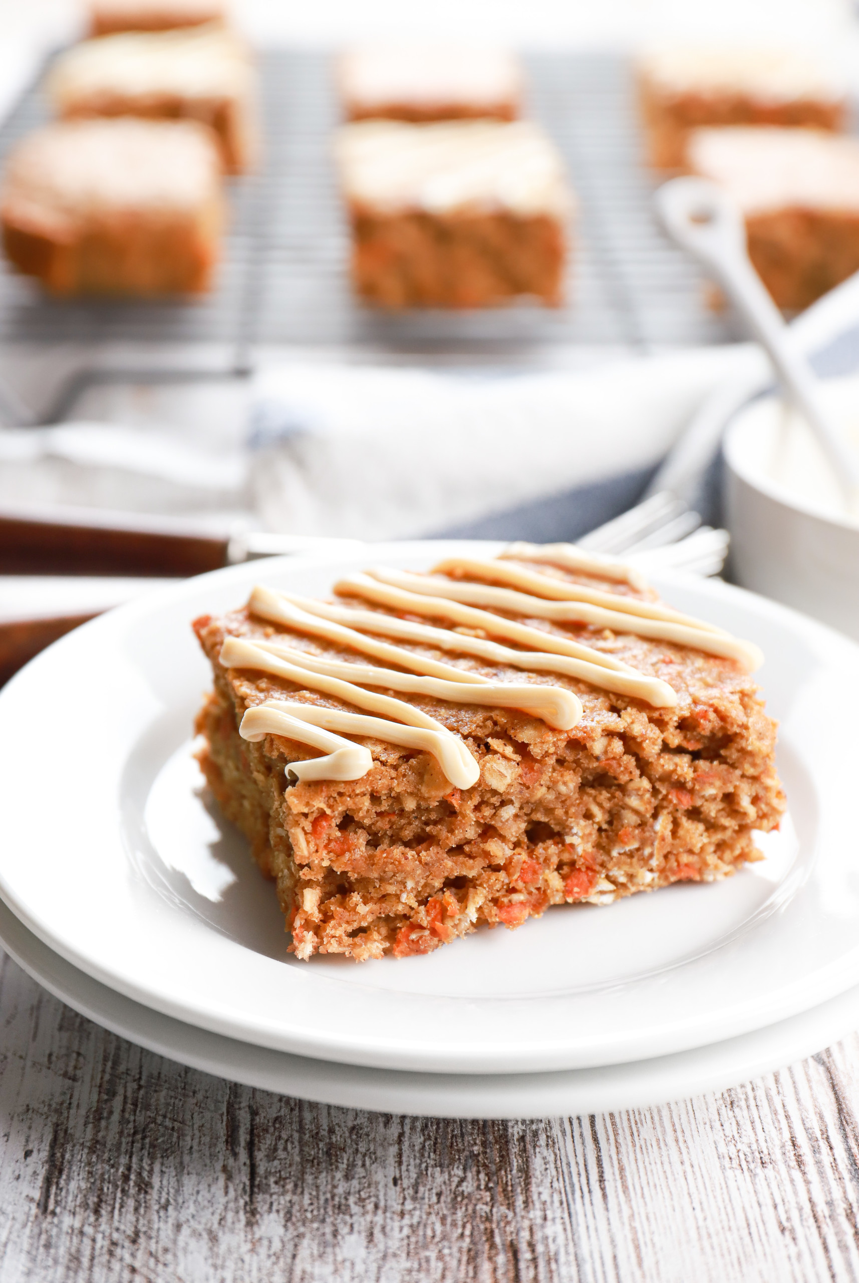 Soft baked carrot cake oat bar on a small white plate with a cooling rack full of bars in the background.