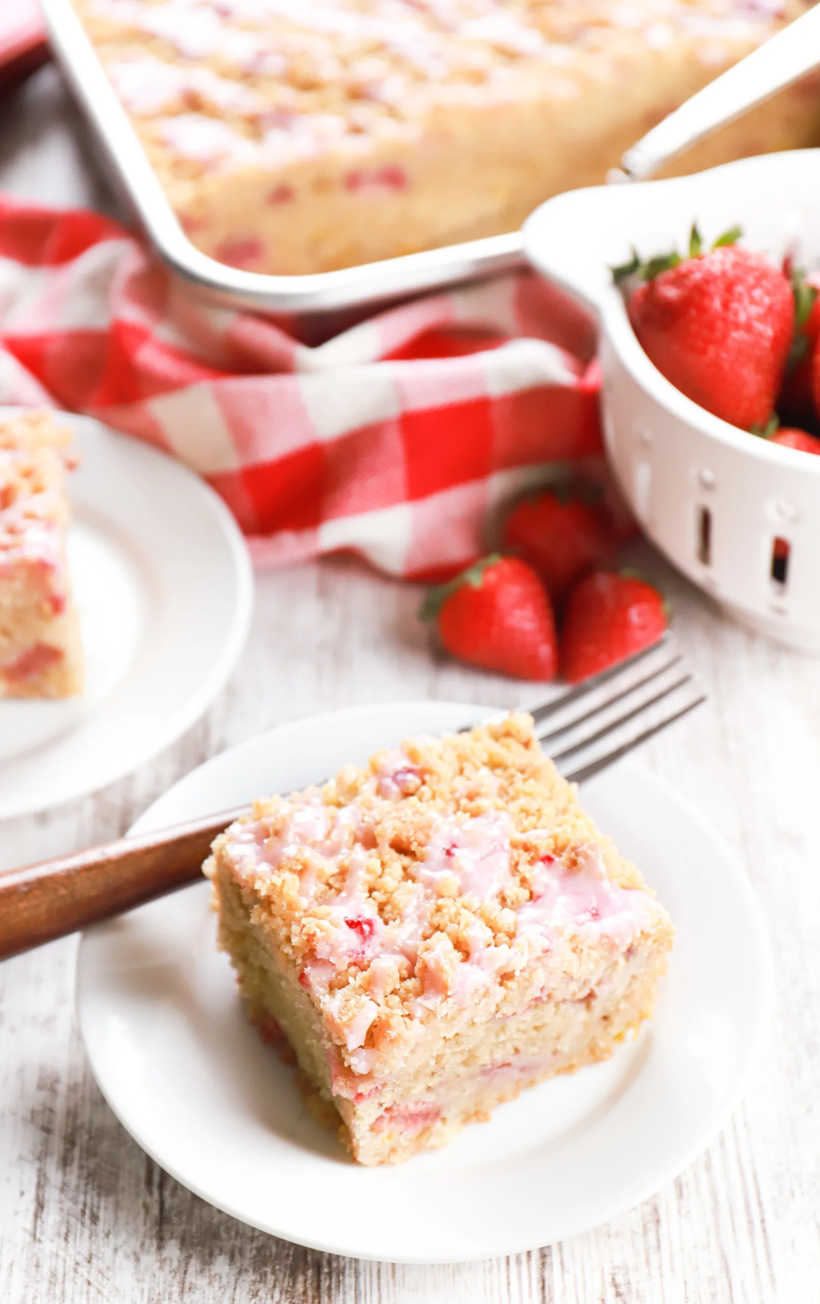 Piece of strawberry rhubarb coffee cake on a small white plate with a small white colander of strawberries and remaining coffee cake in the background.