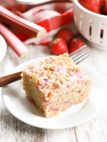One piece of strawberry rhubarb crumb cake on a small white plate with remaining cake in an aluminum cake pan in the background.
