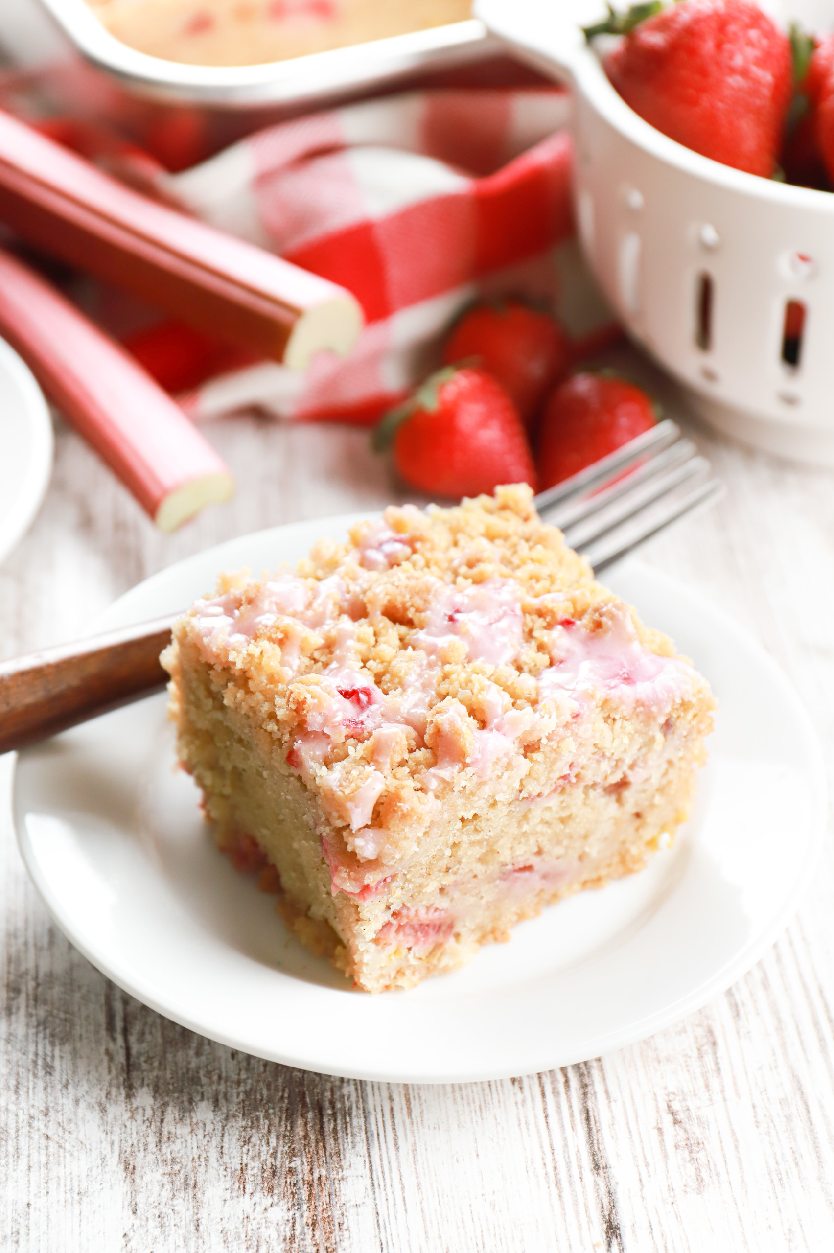 One piece of strawberry rhubarb crumb cake on a small white plate with remaining cake in an aluminum cake pan in the background.