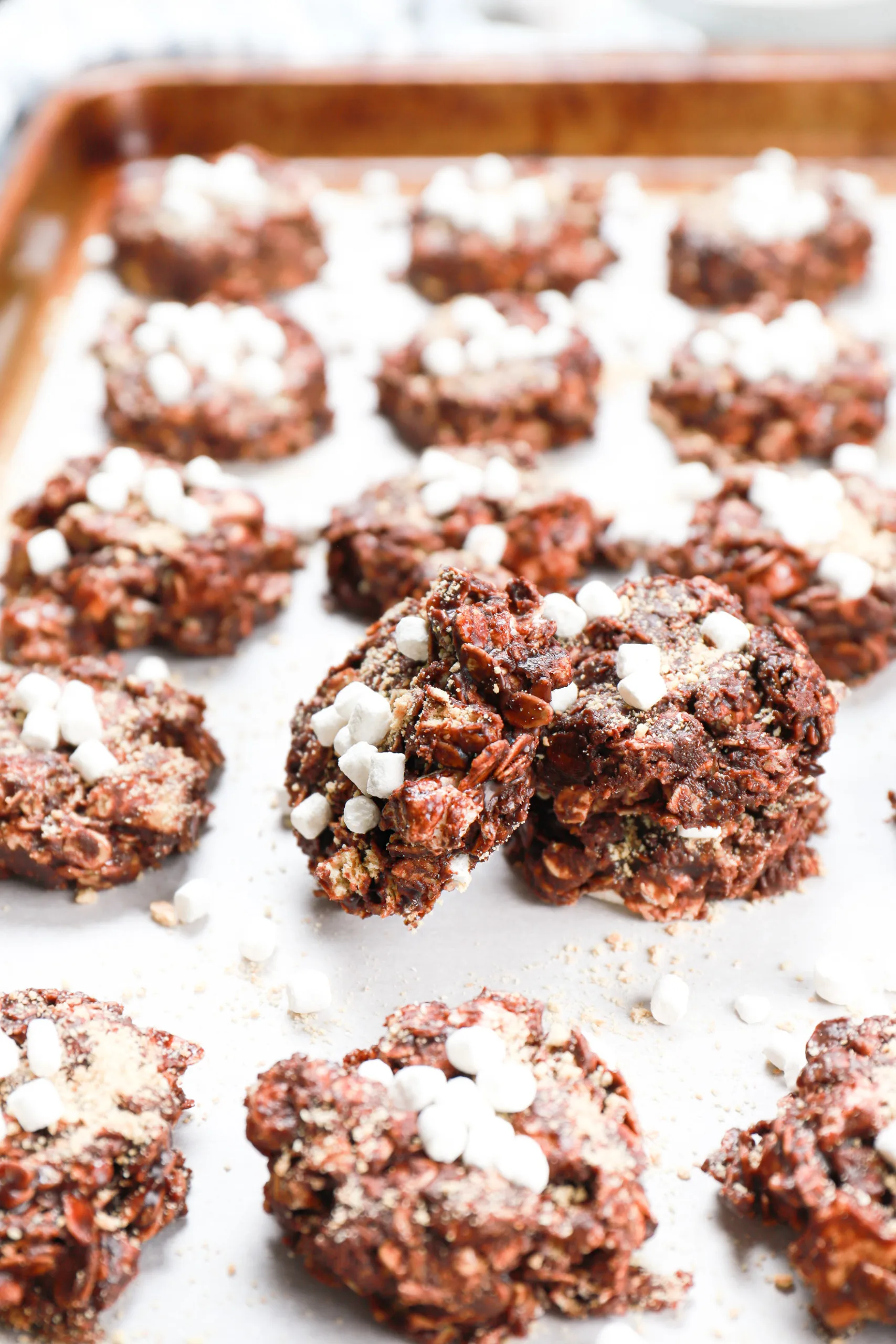 Up close image of a dark chocolate smores no bake cookie leaning against a stack of cookies on a baking sheet with more cookies surrounding it.