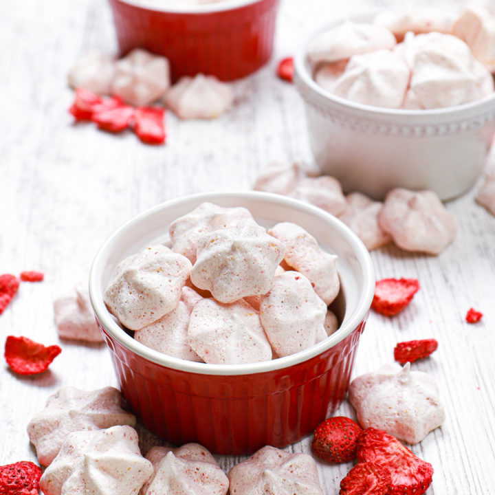 Strawberry lemon mini meringue cookies in a small red bowl surrounded by more cookies and freeze-dried strawberries.