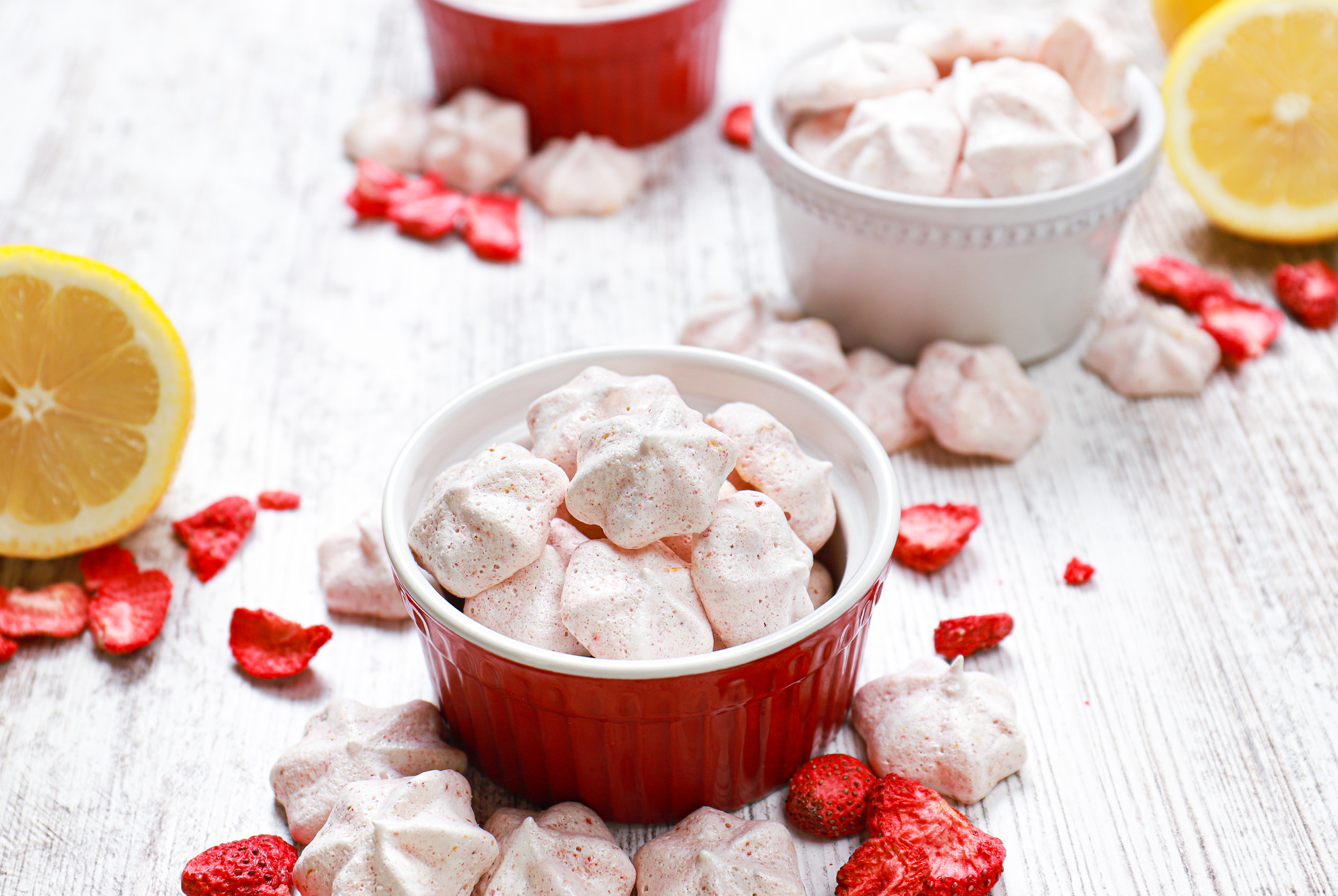 Strawberry lemon mini meringue cookies in a small red bowl surrounded by more cookies and freeze-dried strawberries.