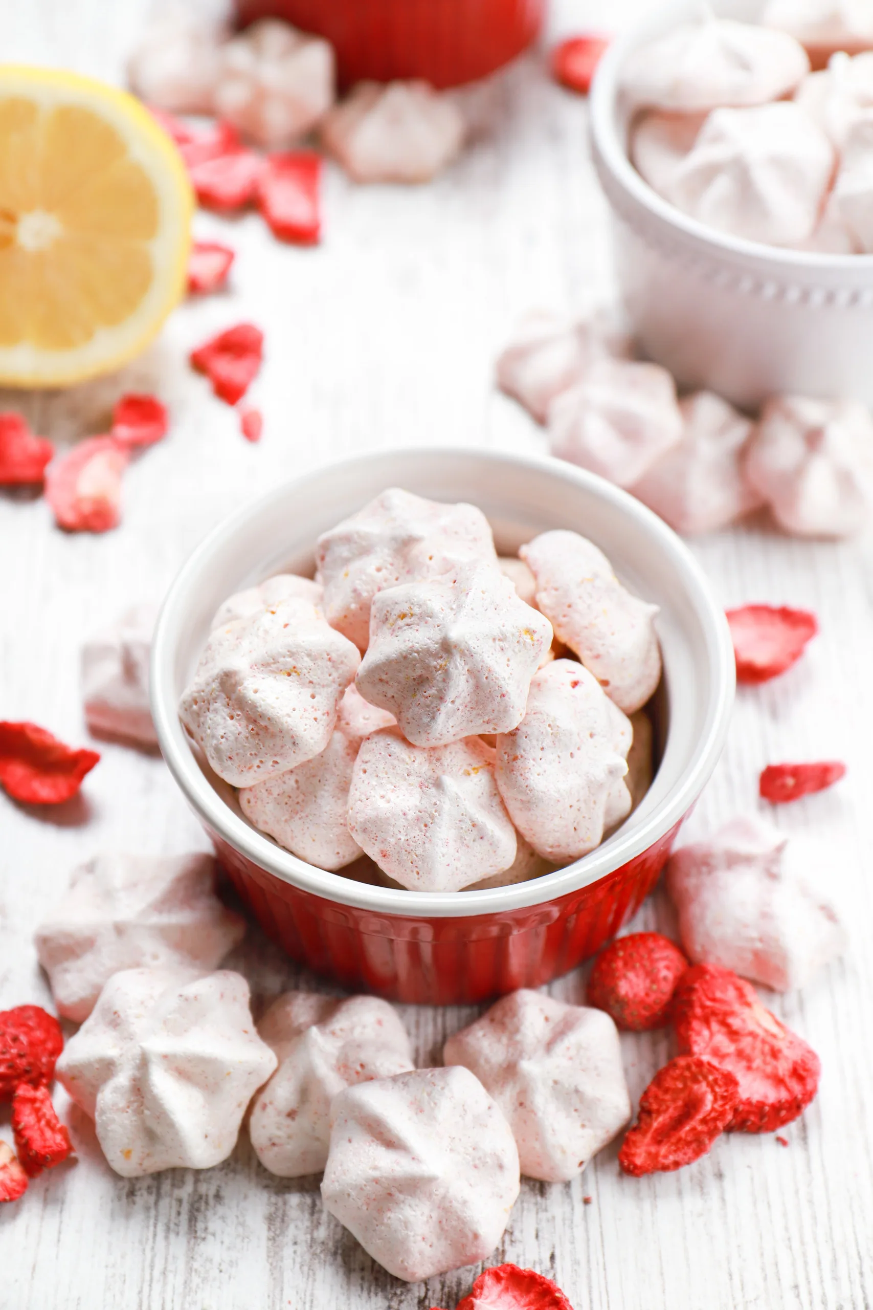 Mini strawberry lemon meringues in small bowls surrounding by a lemon half and more meringues and freeze-dried strawberries.