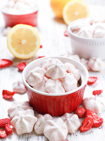 Mini strawberry lemon meringue cookies in small bowls with lemon halves, more meringues, and freeze-dried strawberries surrounding the bowls.