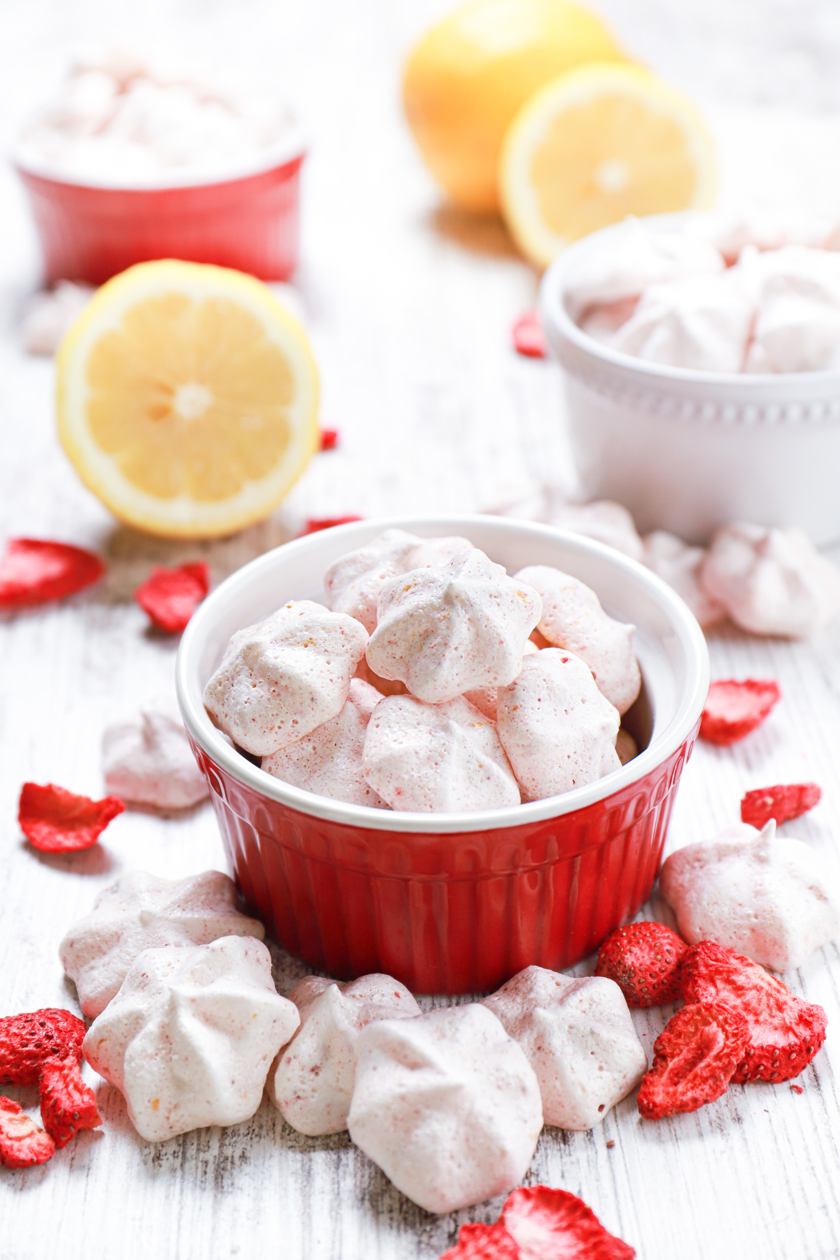 Mini strawberry lemon meringue cookies in small bowls with lemon halves, more meringues, and freeze-dried strawberries surrounding the bowls.