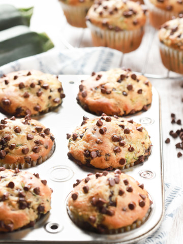 A batch of chocolate chip zucchini muffins in a muffin tin with more muffins on a cooling rack in the background.