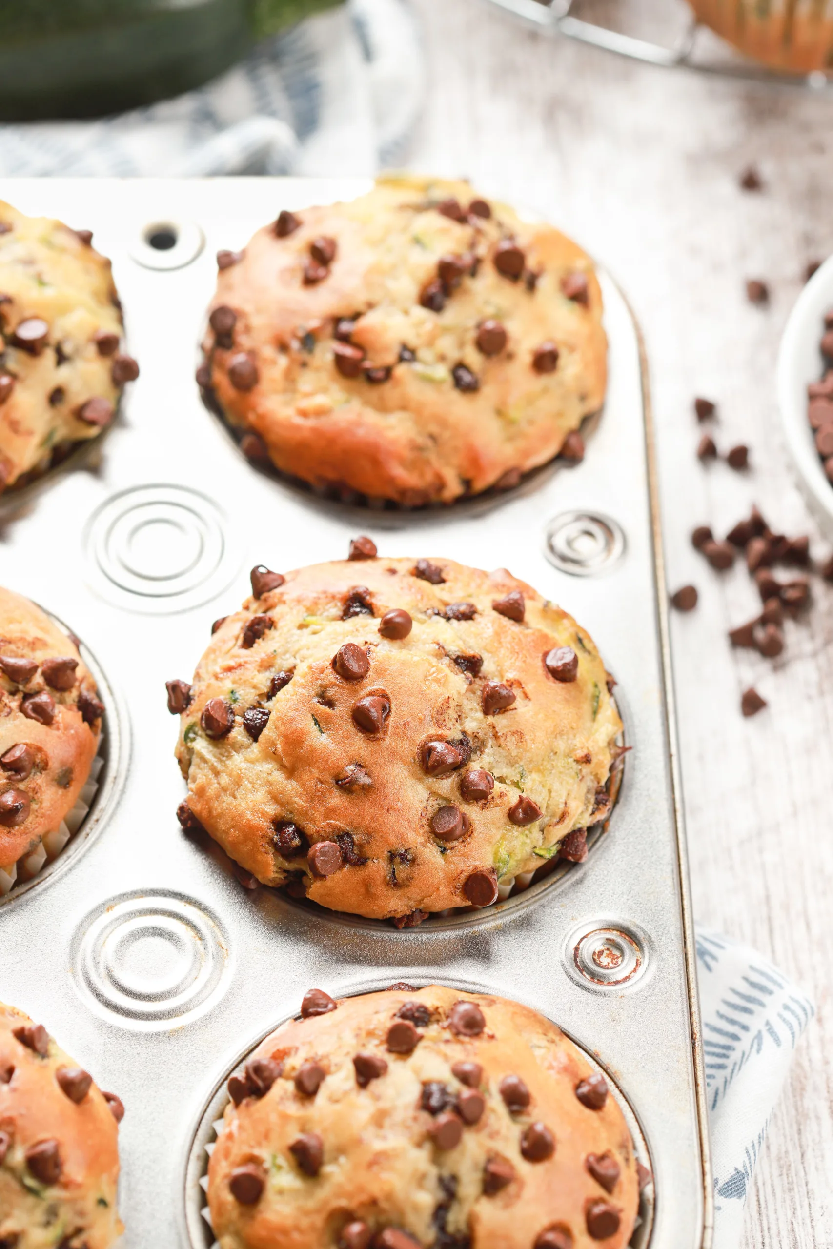 Side view of a batch of bakery style chocolate chip zucchini muffins in a muffin tin on a white wooden board.